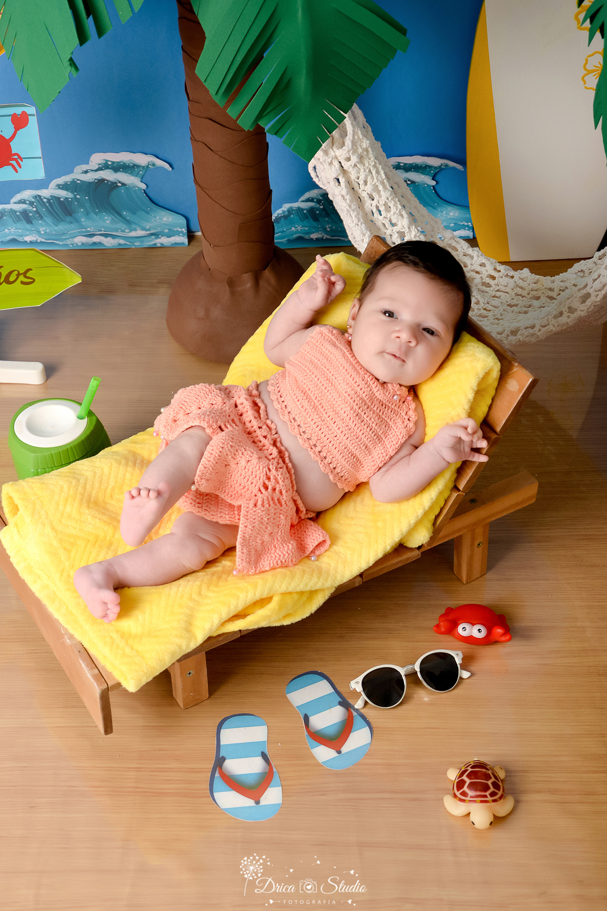 drica studio - ensaio fotográfico infantil - verão e praia - bebe vestinda em roupinha de chochê cor salmão, deitada sobre toalha amarela em espreguiçadeira - carangueijo de brinquedo - coco verde com canudo - piso emitando areia - rede branca