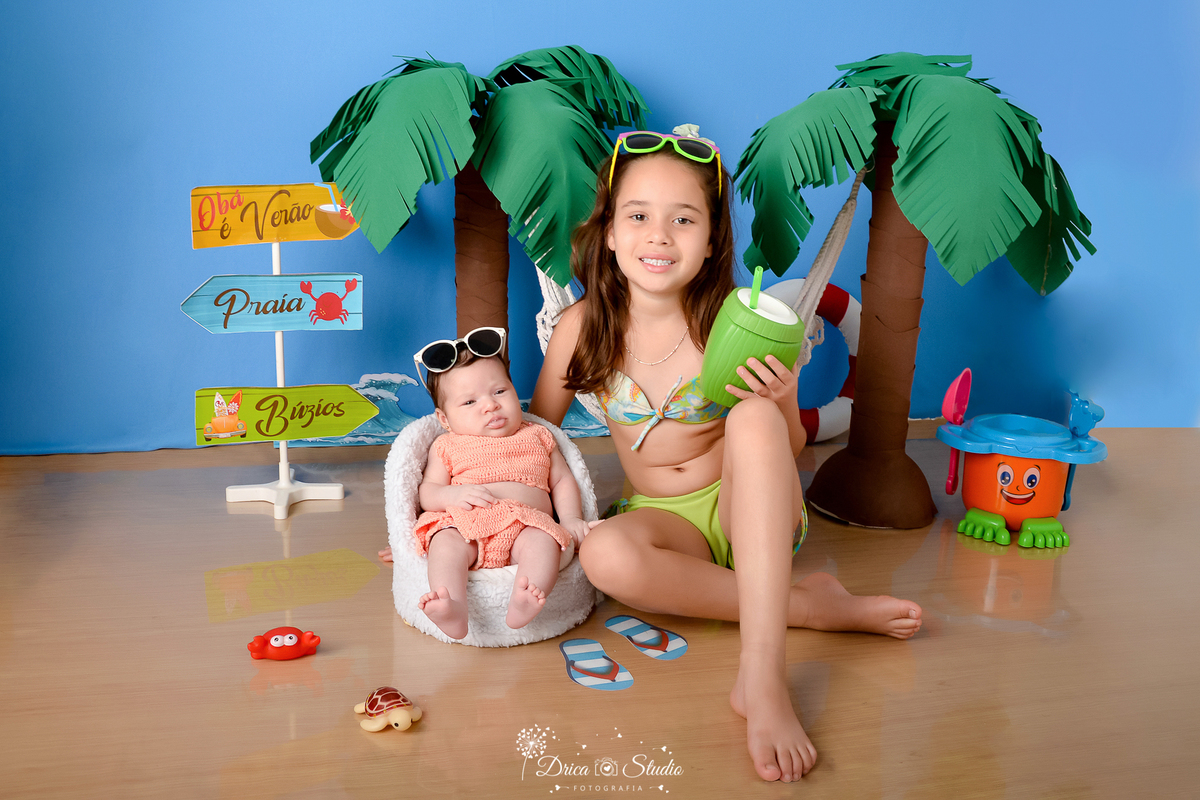drica studio - ensaio fotográfico infantil - verão e praia - bebê vestindo roupa de crochê sendata ao lado da irmã maisl velha que esta vestindo biquini verde segurando um coco verde com canudo - coqueiros - fundo azul - 