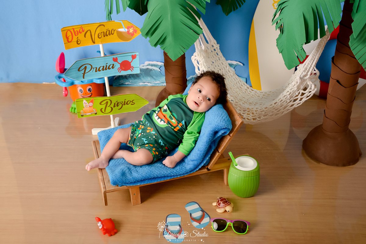 drica studio - ensaio fotográfico infantil - verão e praia - bebê vestindo roupa térmica de praia - sobre manta azul, deitado em espreguiçadeira - rede branca - coqueiros - plaquinhas de indicação - piso emitando areia 