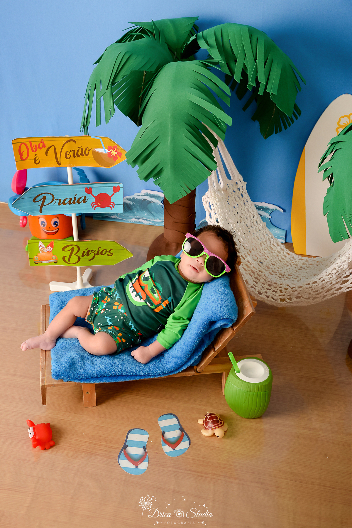 drica studio - ensaio fotográfico infantil - verão e praia - bebê vestindo roupa termica de sol, usando óculos escuro - rede branca - plaquinha de indicação - fundo azul - onda do mar - coqueiro - coco verde com canudo