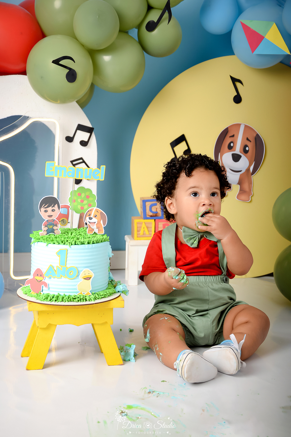 drica studio - ensaio fotográfico infantil - smash the cake - bento e o toto - bebê sentado ao lado do bolo com tema bento e o toto - painel amarelo - notas musicais - camisa vermelha e short verde claro