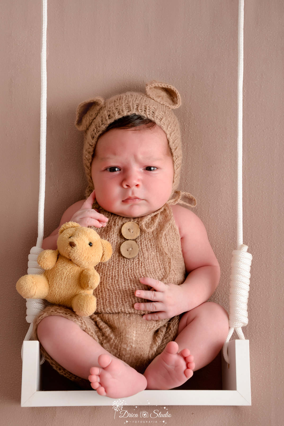 dricastudio - newborn - ensaiofotograficoinfantil - xinguara - bebê sentado em pequeno balanço branco, usando um conjunto de tricô bege com botões frontais, segurando um ursinho de pelúcia amarelo - fundo bege