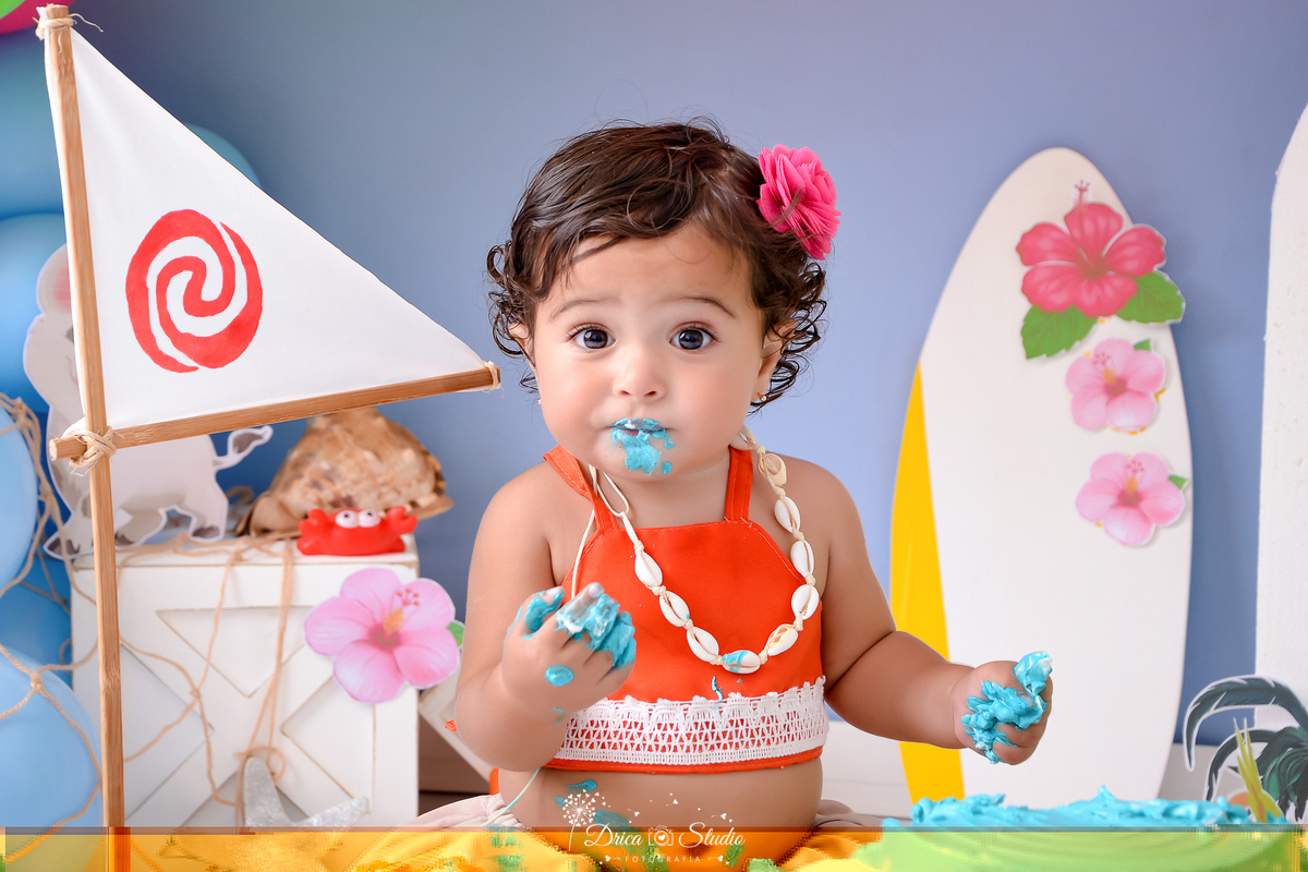 drica studio - ensaio fotografico infantil - smash the cake moana baby - xinguara - bebê vestido top laranja e saia bege, usando flor rosa no cabelo, com as mãos e a boca suja de cobertura azul de bolo - pracha branca - caixote branco - concha - fundo azu
