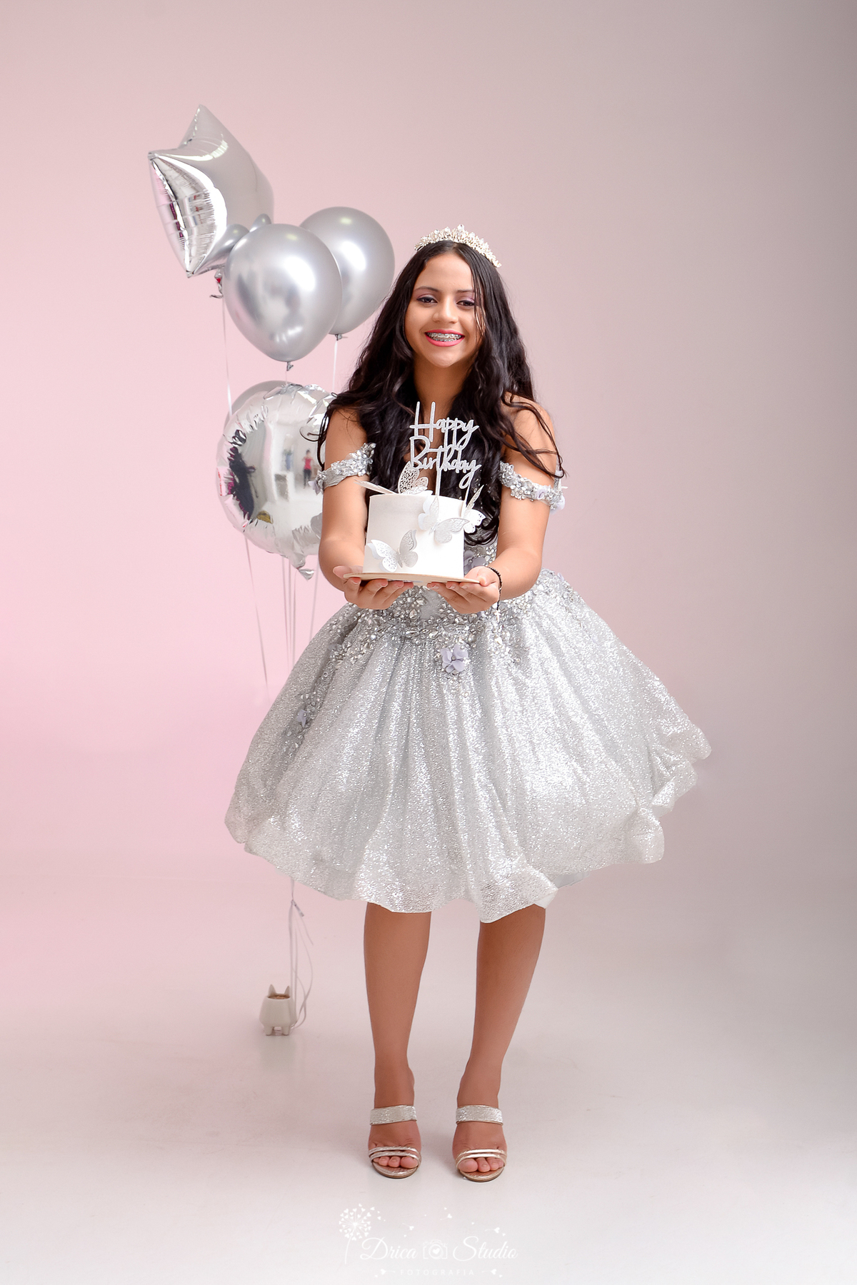 rica studio - ensaio fotográfico 15 anos - xinguara - jovem segura à frente do corpo bolo branco - vestido prateado curto estilo princesa, na cabeça tiara de brilhante sobre a cabeça - balão prata - fundo rosado