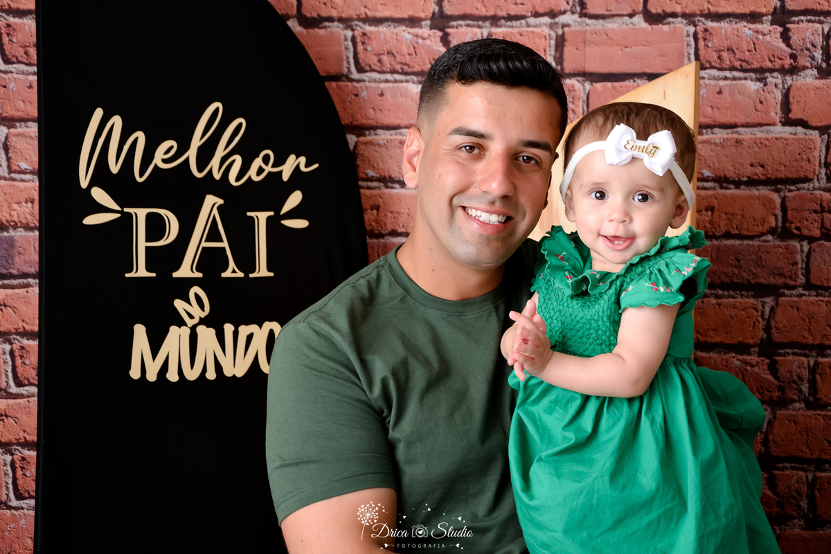 drica studio - ensaio fotografico dia dos pais - xinguara - Pai vestindo camisa verde militar segurando bebê no colo - bebê usando vestido verde com laço branco na cabeça - fundo de tijolos vermelho - painel preto com frase 