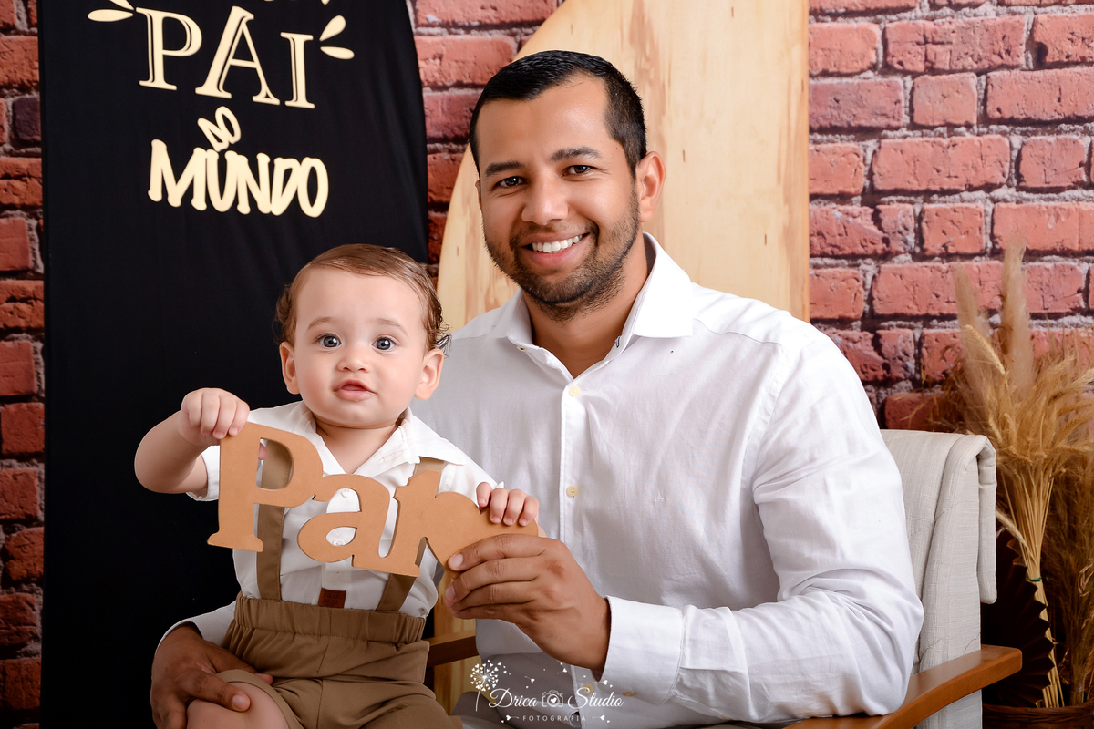 drica studio - ensaio fotografico dia dos pais - xinguara - pai sentado em poltrona cinza com detalhes de madeira, com bebê no colo, ambos usando camisa branca com calça bege - fundo de tijolos vermelho e painel preto com frase
