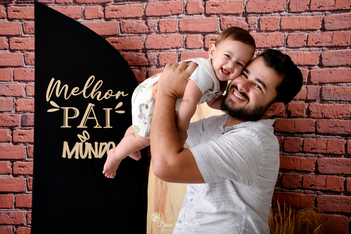 drica studio - ensaio fotografico dia dos pais - xinguara - pai veste camisa branca, segurando bebê no ar, próximo ao seu rosto - bebê veste macaquinho com desenho de bichinhos e camisa branca - fundo de tijolos vermelho - painel preto.