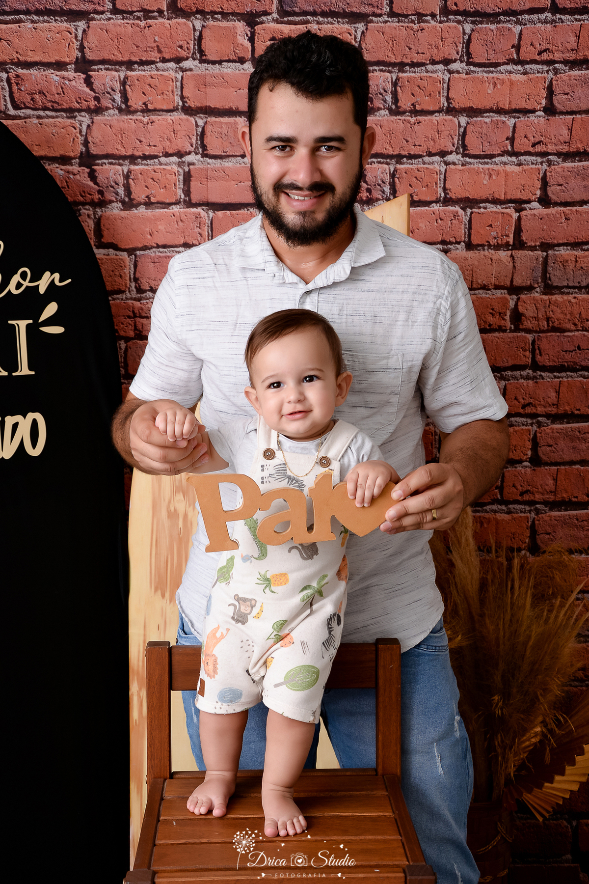 drica studio - ensaio fotografico dia dos pais - xinguara -pai veste camisa branca e segura o filho em pé em cima de uma cadeira - bebê  segura plaquinha com nome pai - fundo de tijolos vermelho - painel preto com frase 