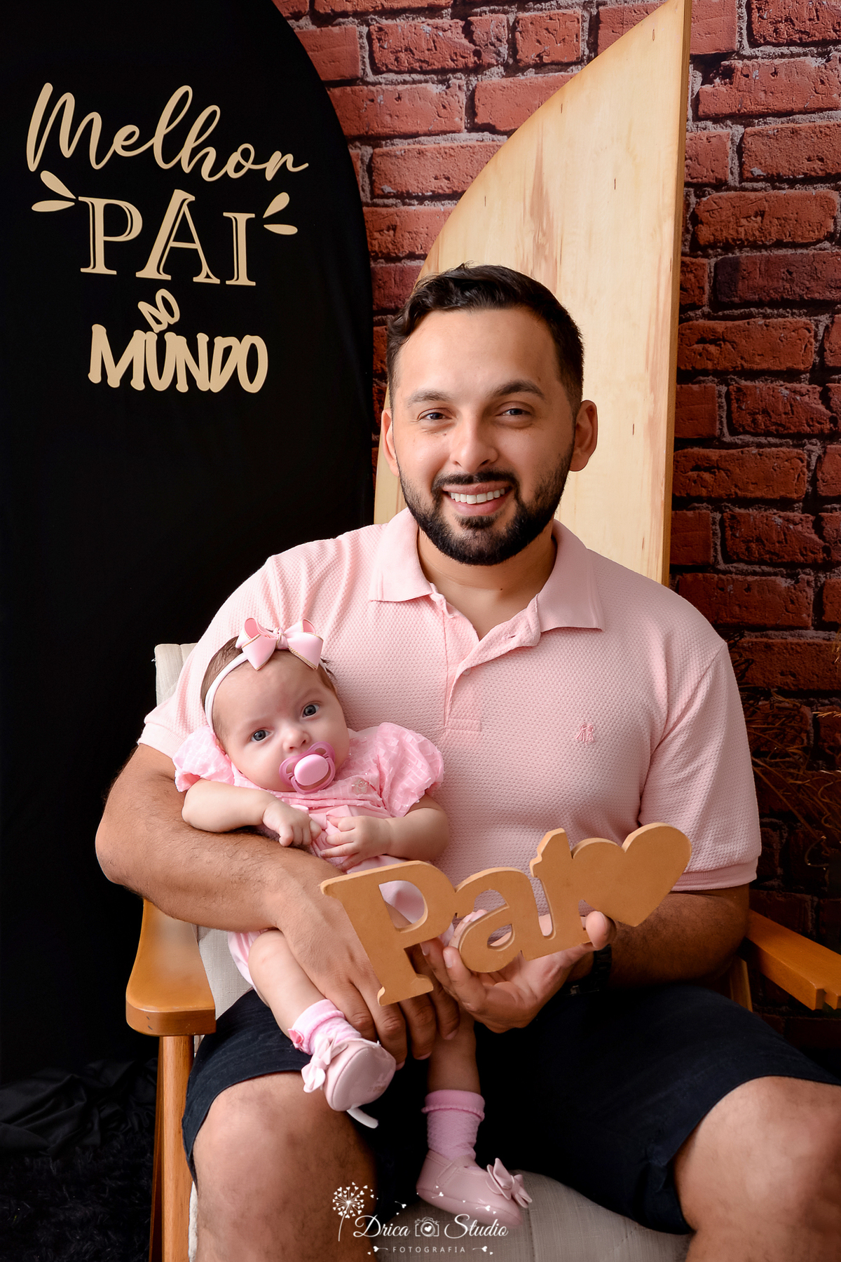 drica studio - ensaio fotografico dia dos pais - xinguara - vai segurando filha bebê no colo - camisa rosa claro - vestido e laço rosa claro - fundo de tijolos vermelho - painel preto com frase 