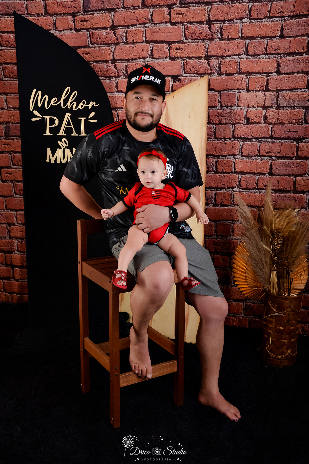 drica studio - ensaio fotografico dia dos pais - xinguara - pai usando camisa do flamento segurando filha no coloco, também vestindo bory vermelho com brasão do flamengo - fundo de tijolos vermelho- painel preto com frase