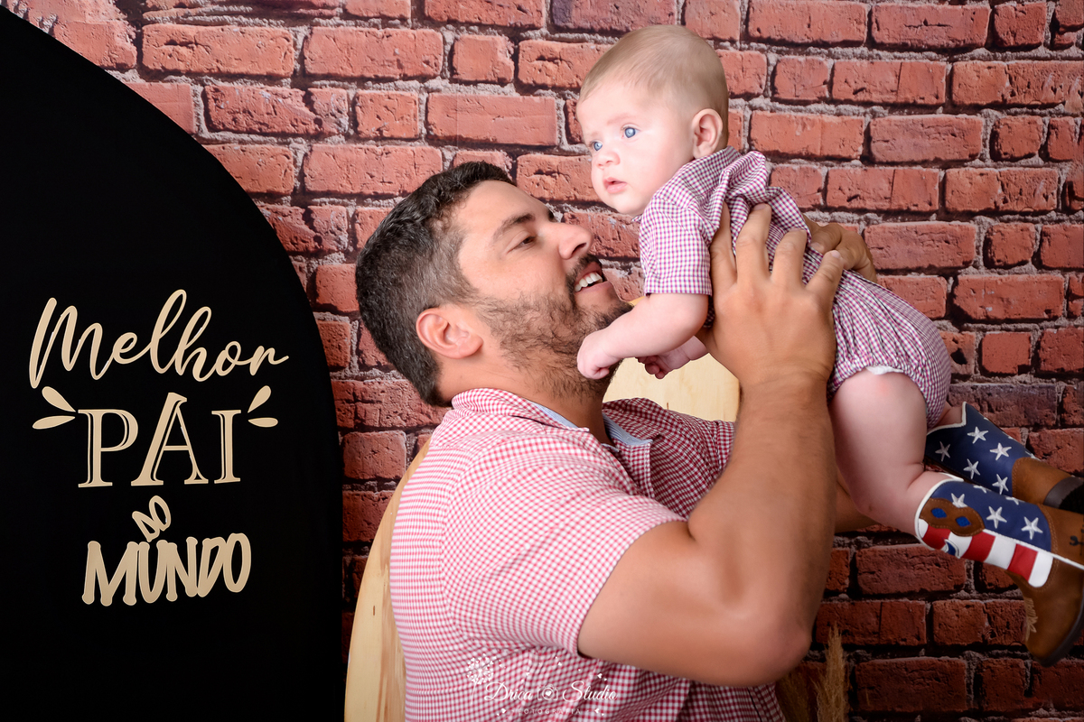 drica studio - ensaio fotografico dia dos pais - xinguara - pai usando camisa xadrez vermelha, segurando filho no ar, bebê veste bory xadres vermelho, calça bota com detalhes da bandeira americana - fundo de tijolos vermelho - painel preto