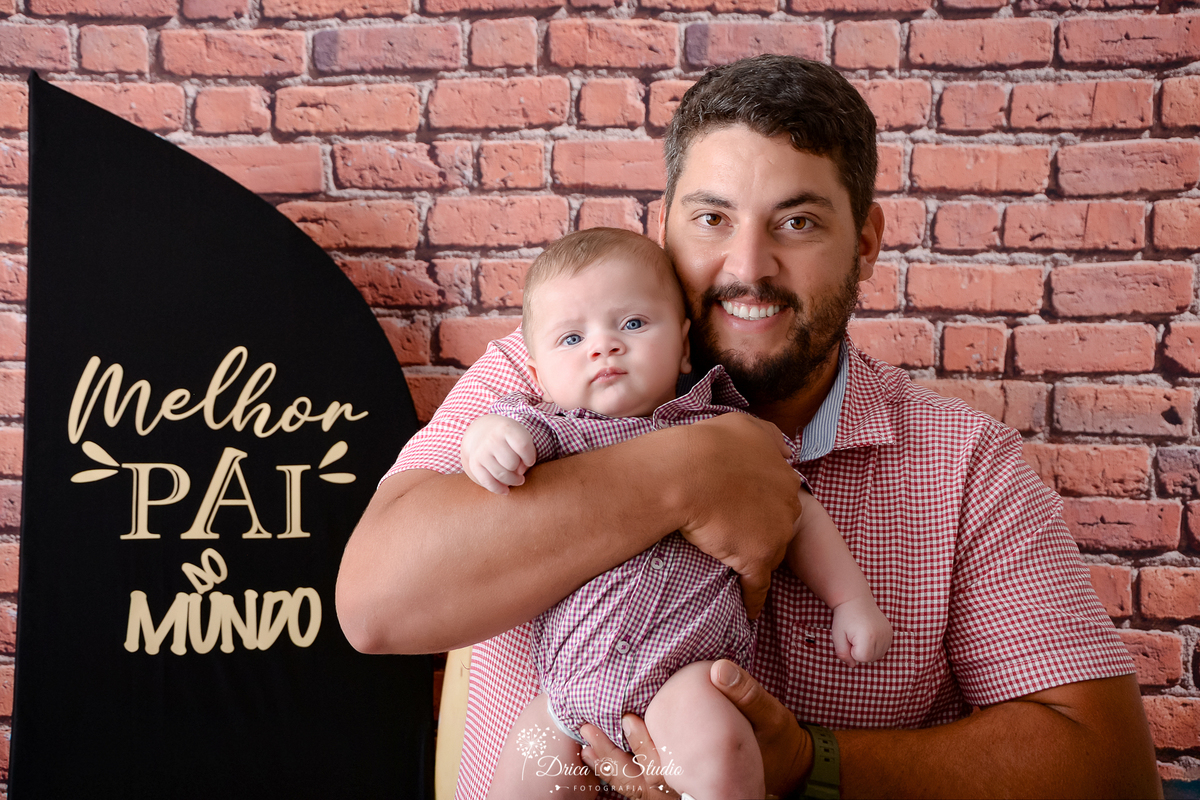 drica studio - ensaio fotografico dia dos pais - xinguara - pai veste camisa xadrez vermelha e segura bebê a sua frente - bory xadrez vermelho  - fundo de tijolos vermelho - painel preto com frase 