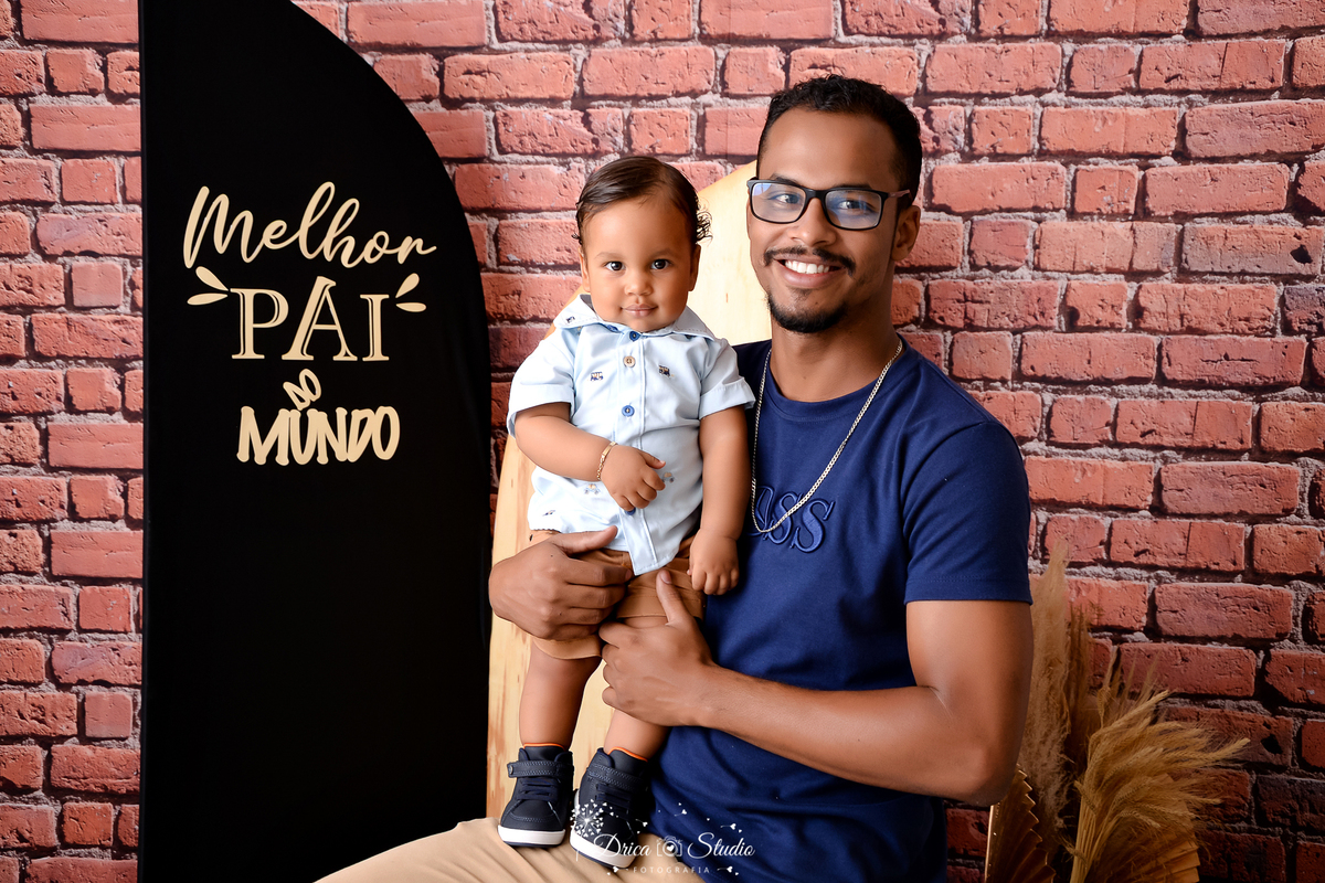 drica studio - ensaio fotografico dia dos pais - xinguara - pai usando camisa azul, seguando filho em pé sobre sua perna- bebê vesti camisa azul clara - fundo de tijolos vermelho - painel preto com frase 