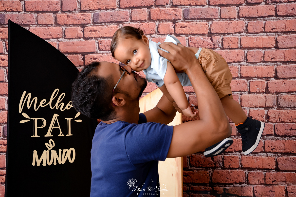 drica studio - ensaio fotografico dia dos pais - xinguara - pai segurando filho no ar, proximo ao seu rosto, vestindo camisa azul escura - bebê usando camisa azul clara e short marrom - tenis preto - fundo de tijolos vermelho e painel preto