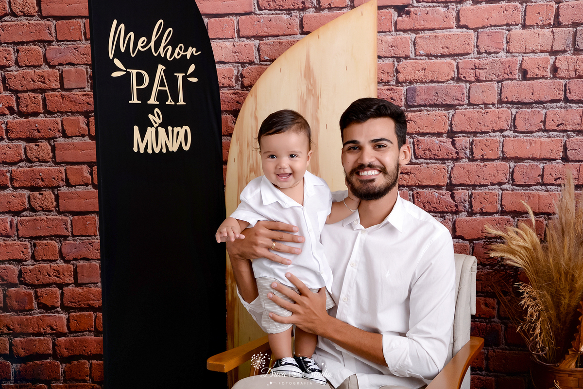 drica studio - ensaio fotografico dia dos pais - xinguara - pai sentando em poltrona, segurando filho em pé em seu colo, ambos usando camisa branca - short bege e calça bege - folhagem seca - fundo de tijolos vermelho - painel preto 