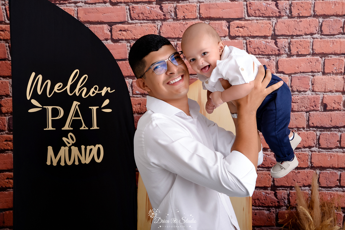 drica studio - ensaio fotografico dia dos pais - xinguara - pai segura bebê no ar próximo ao rosto, ambos veste camisa branca e calça azul - folhagem seca - fundo de tijolos vermelho - painel preto com frase 