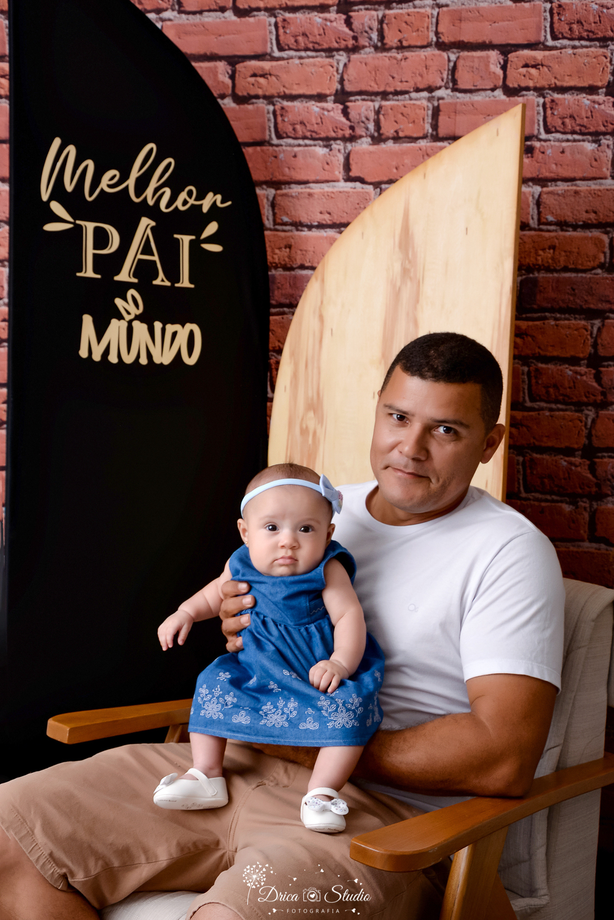 drica studio - ensaio fotografico dia dos pais - xinguara - pai segurando filha no colo - camisa branca - vestido azul escuro - laço azul claro - fundo de tijolos vermelho - painel preto e madeirado com frase 