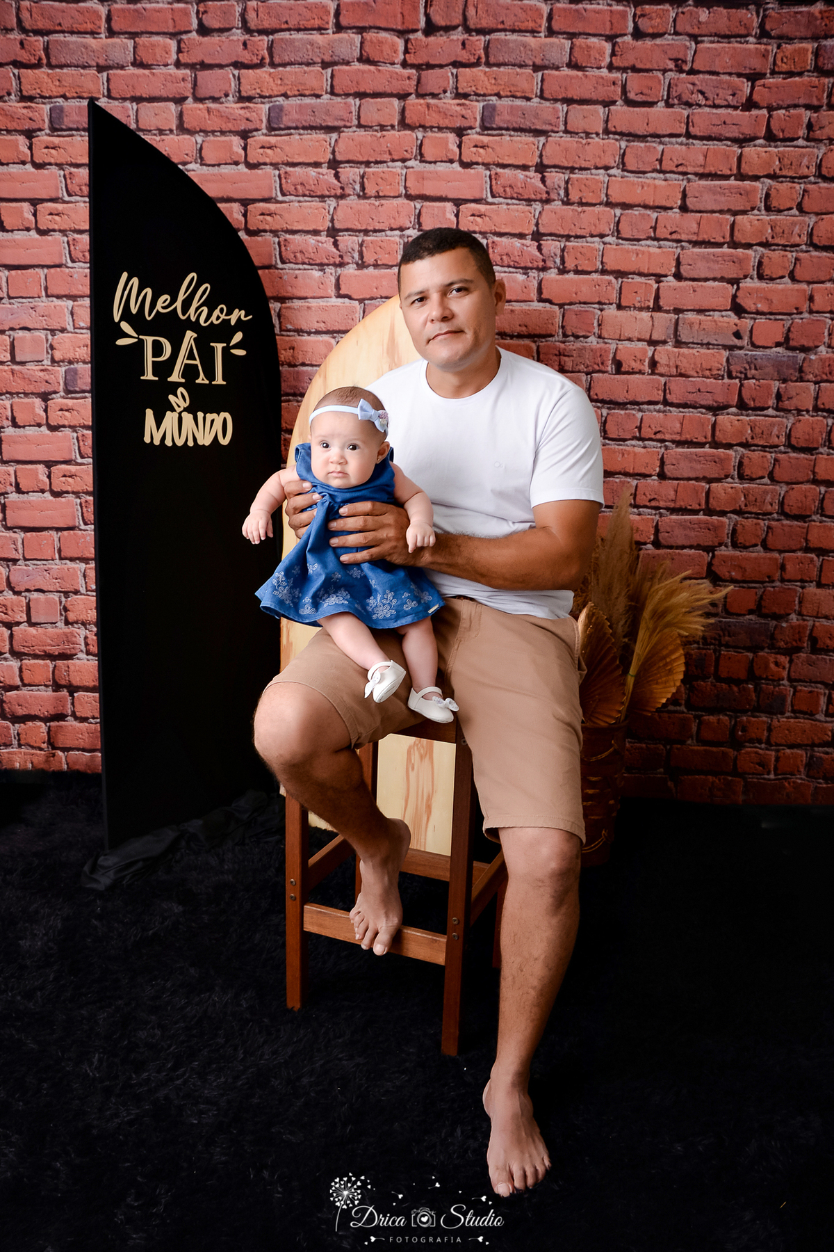 drica studio - ensaio fotografico dia dos pais - xinguara - homem sentado em cadeira com filha no colo - camisa branca - short bege escuro - vestido azul escuro - laço azul claro - saparinho branco - fundo de tijolos vermelho - painel preto