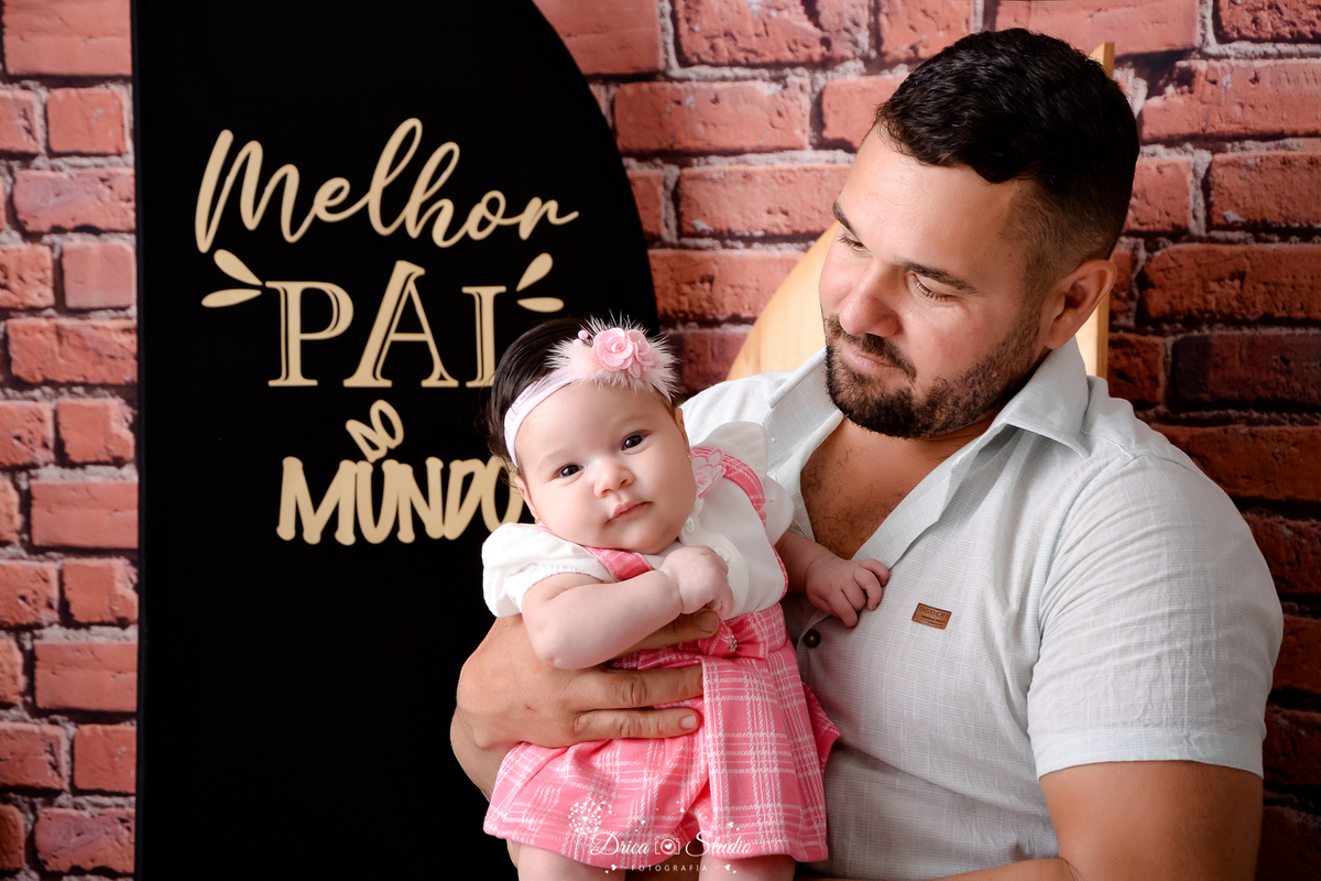 drica studio - ensaio fotografico dia dos pais - xinguara - homem vestindo camisa branca segurando bebê, que veste jardineira rosa com blusa branca - tiara rosa - fundo de  tijolos vermelho e painel preto com frase
