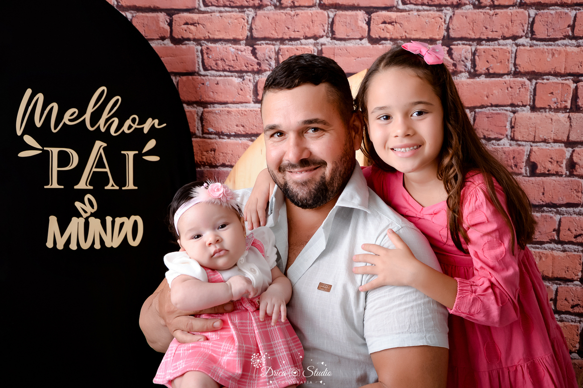 drica studio - ensaio fotografico dia dos pais - xinguara - pai segurando bebê no colo e filha mais velha em pé atrás dele - camisa branca - rardineira rosa com blusa banca - vestido de manga longa rosa - fundo de tijolos vermelho - painel preto
