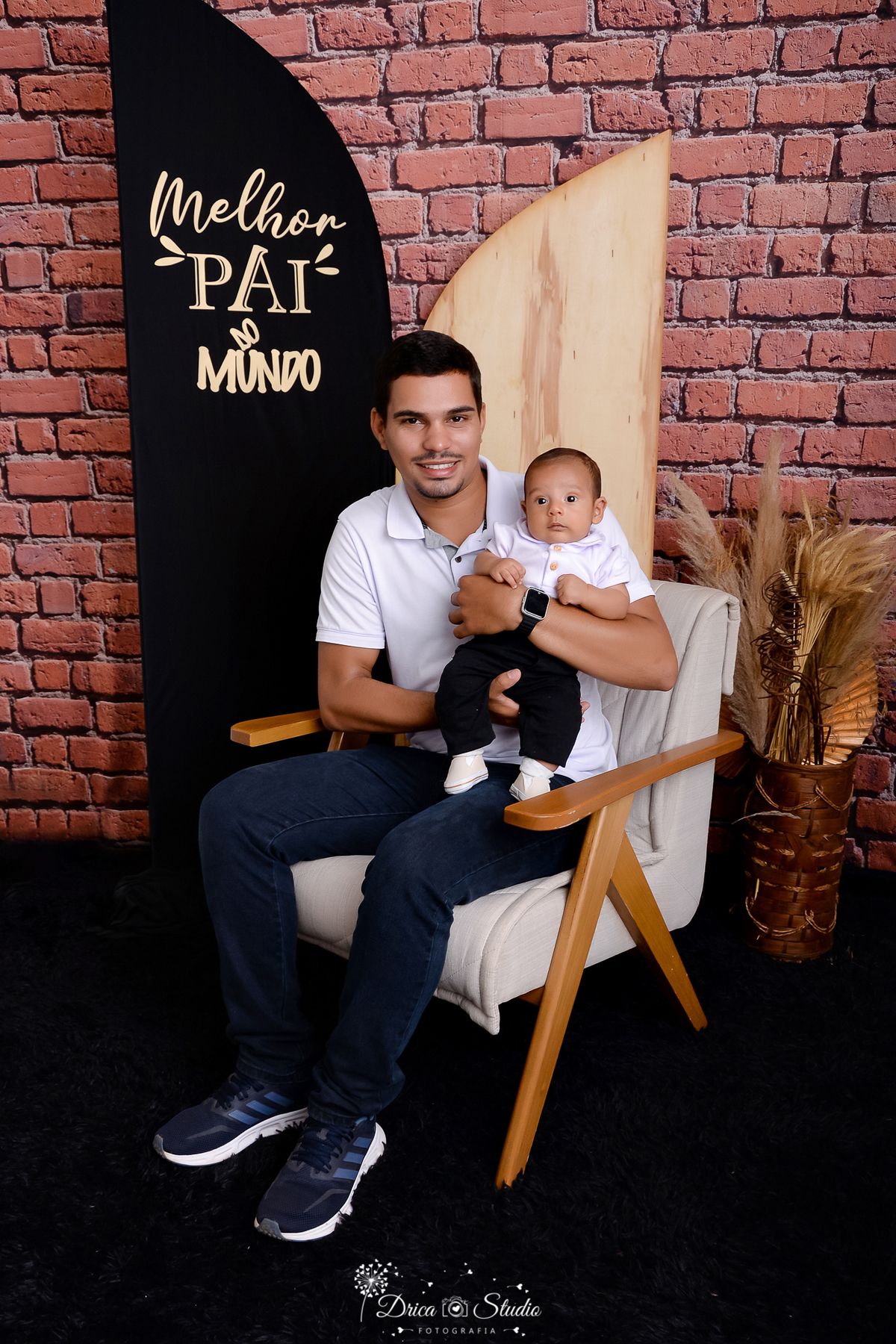 drica studio - ensaio fotografico dia dos pais - xinguara - homem sentado em poltrona bege com detalhe em madeira segurando bebê no colo, ambos vestindo camisa branca e calça jeans azul - fundo de tijolos vermelho - painel preto - folhagem seca