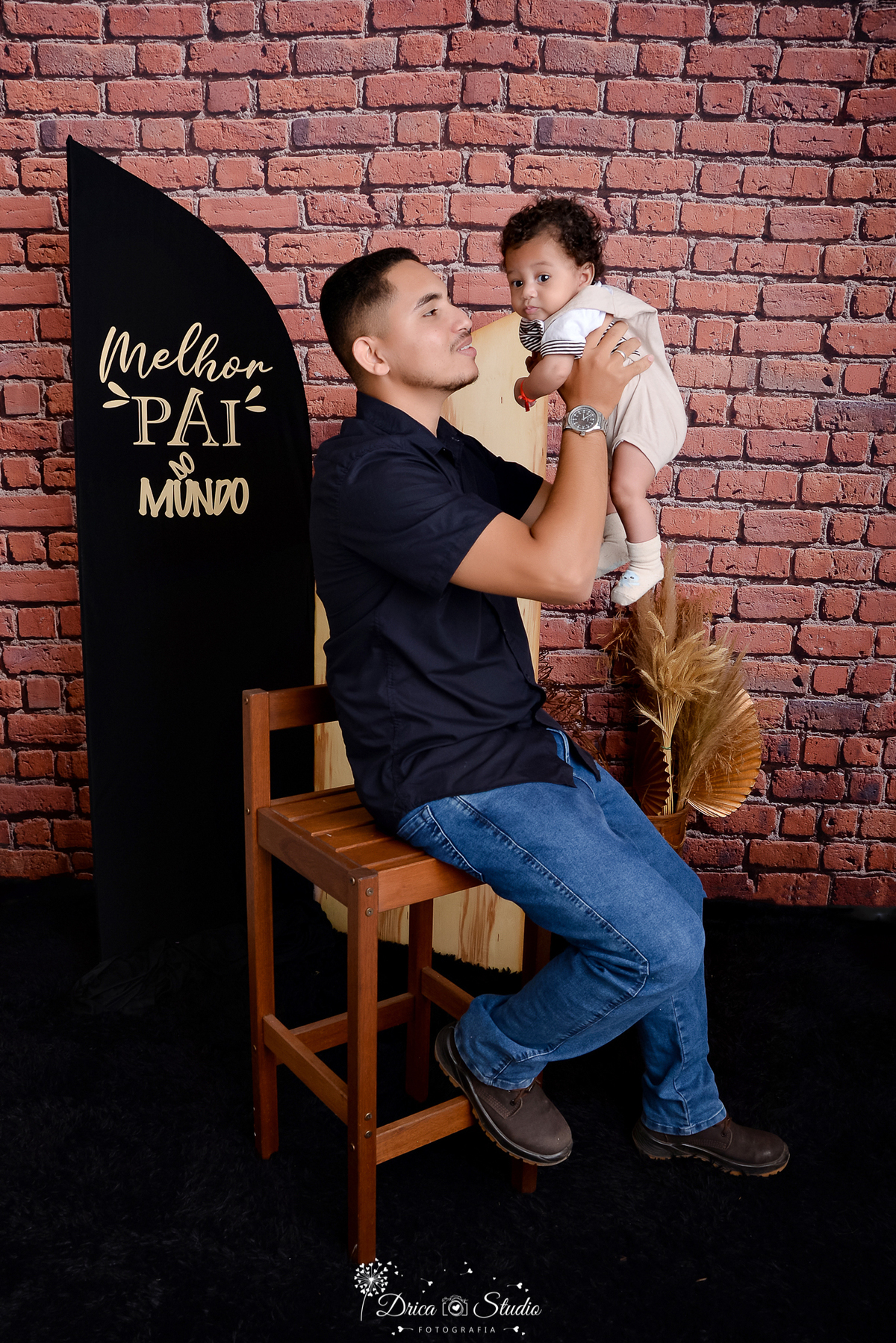 drica studio - ensaio fotografico dia dos pais - xinguara - homem sentando em cadeira, segurando filho no ar próximo ao rosto - camisa preta - calça jeans azul - macaquinho bege - fundo de tijolos vermelho e painel preto com frase 