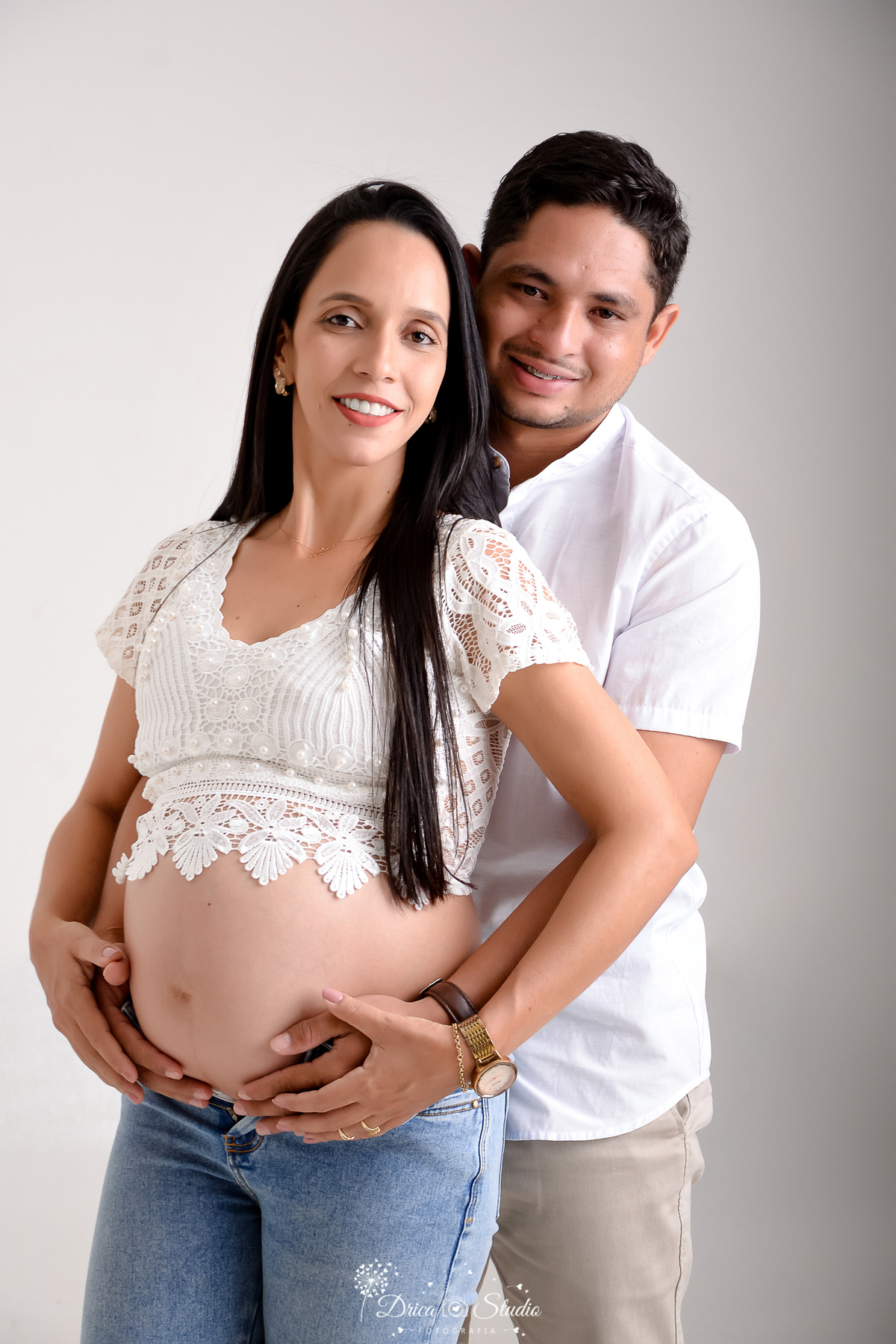 drica studio - ensaio fotográfico gestante - xinguara - mulher gestante usando blusa branca com renda delica e calça jeans - homem posionado atras da mulher vestindo camisa branca e calça jeans, envolvendo com os braços e apoiando as mão sobre barriga