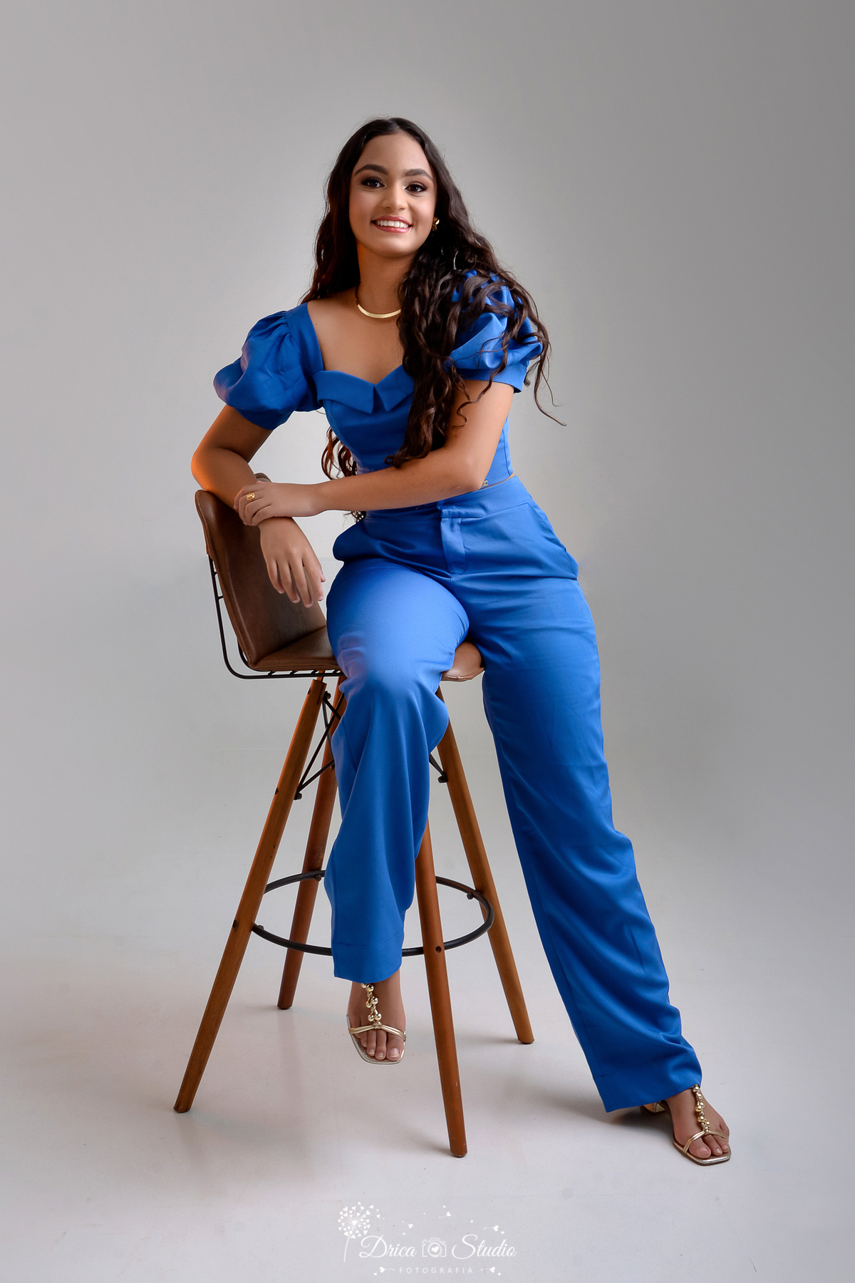 drica studio - ensaio fotográfico 15 anos - xinguara - jovem de cabelos longos e cacheados sentada em cadeira alta de couro e madeira, usando croped e calça de alfaiataria azul - colar dourado - fundo cinza claro