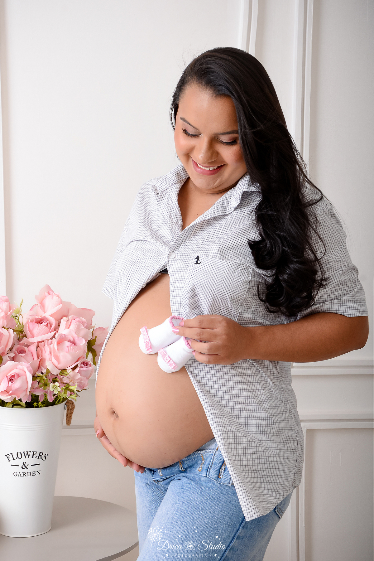 drica studio - ensaio fotográfico - gestante - gestante usado camisa aberta na frente com barriga a amostra, segurando um sapatinho sobre a barriga - rosa cor de rosa - fundo branco com boiserie 