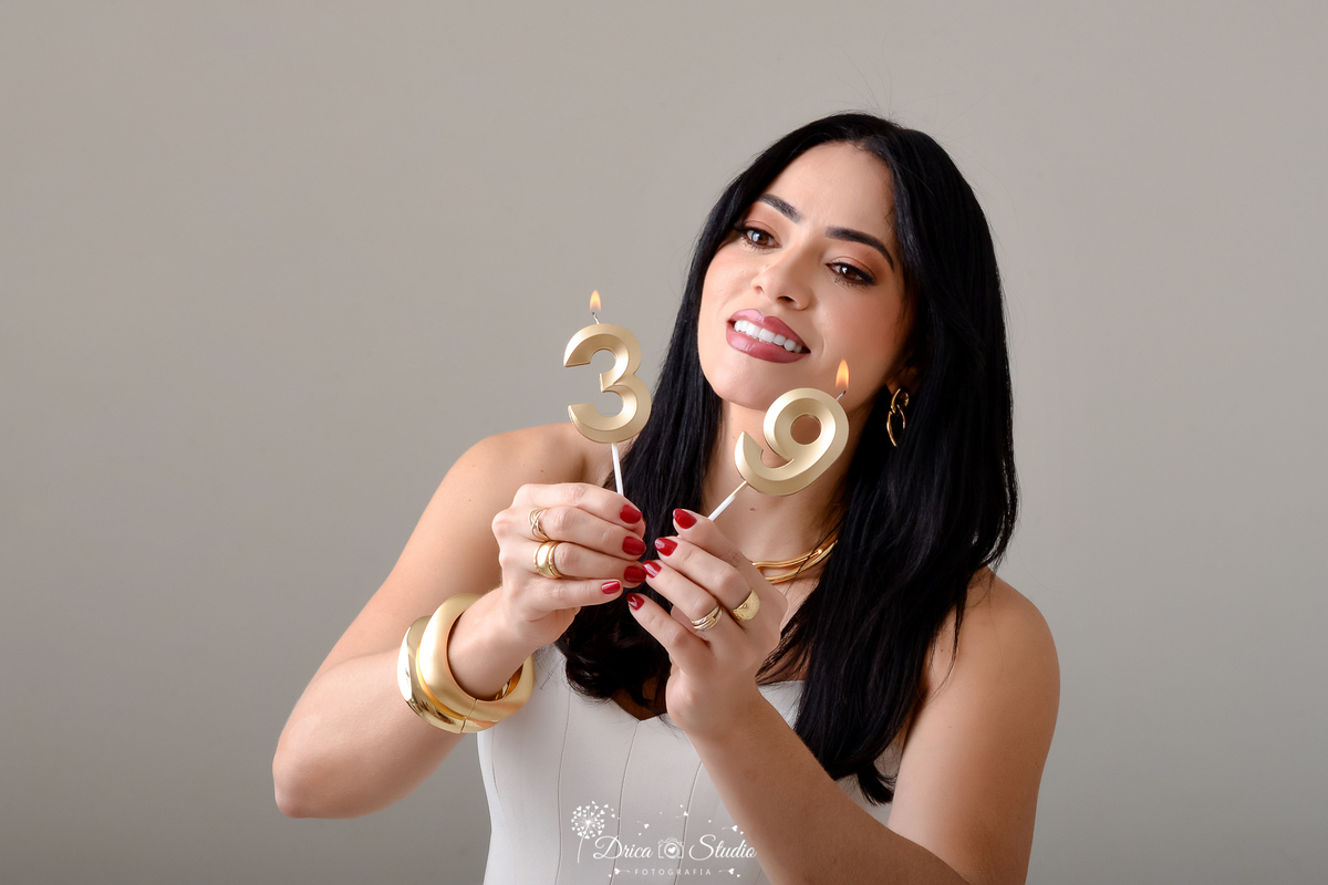 drica studio - ensaio fotografico - aniversário - profissional - xinguara - mulher de cabelos escuro vestindo croped na cor creme estruturado, segurando velinhas dourada com numero trinta e nove - braceletes dourados - fundo neutro