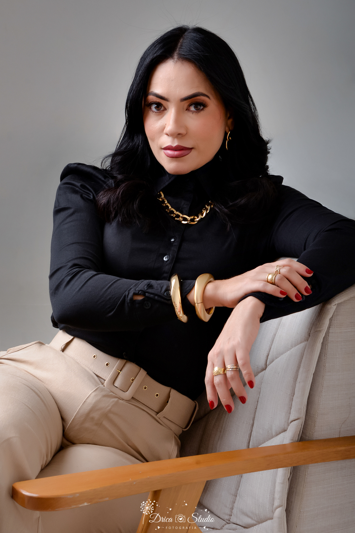 drica studio - ensaio fotografico - aniversário - profissional - xinguara - mulher sentada em poltrona bege, vestindo camisa preta de manga longa e calça de alfaiataria bege, usa bracelete grosso e colar de estilo corrente dourado - fundo neutro
