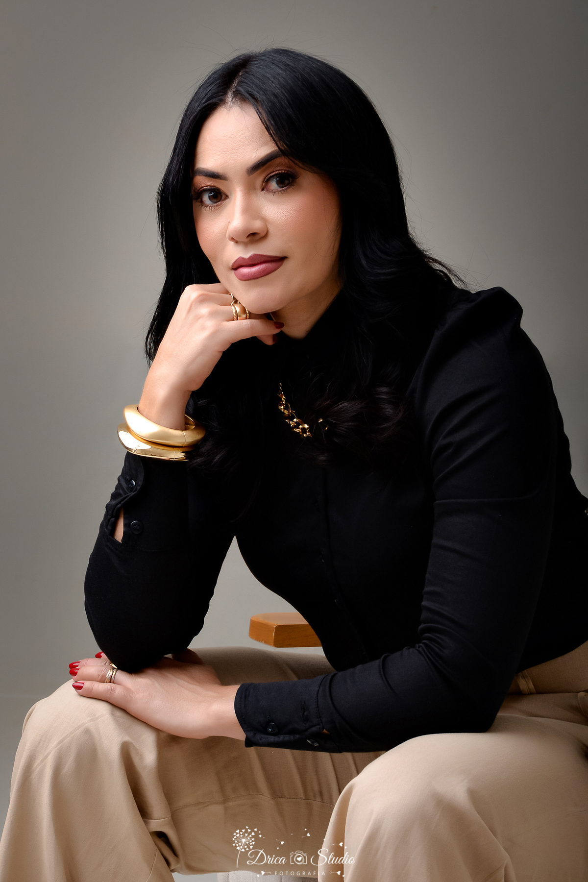 drica studio - ensaio fotografico - aniversário - profissional - xinguara - mulher sentada com corpo levente inclado para frente e apoiado sobre as pernas - usando camisa preta de manga longa e calça bege -braceletes grosso dourado e colar estilo corrente