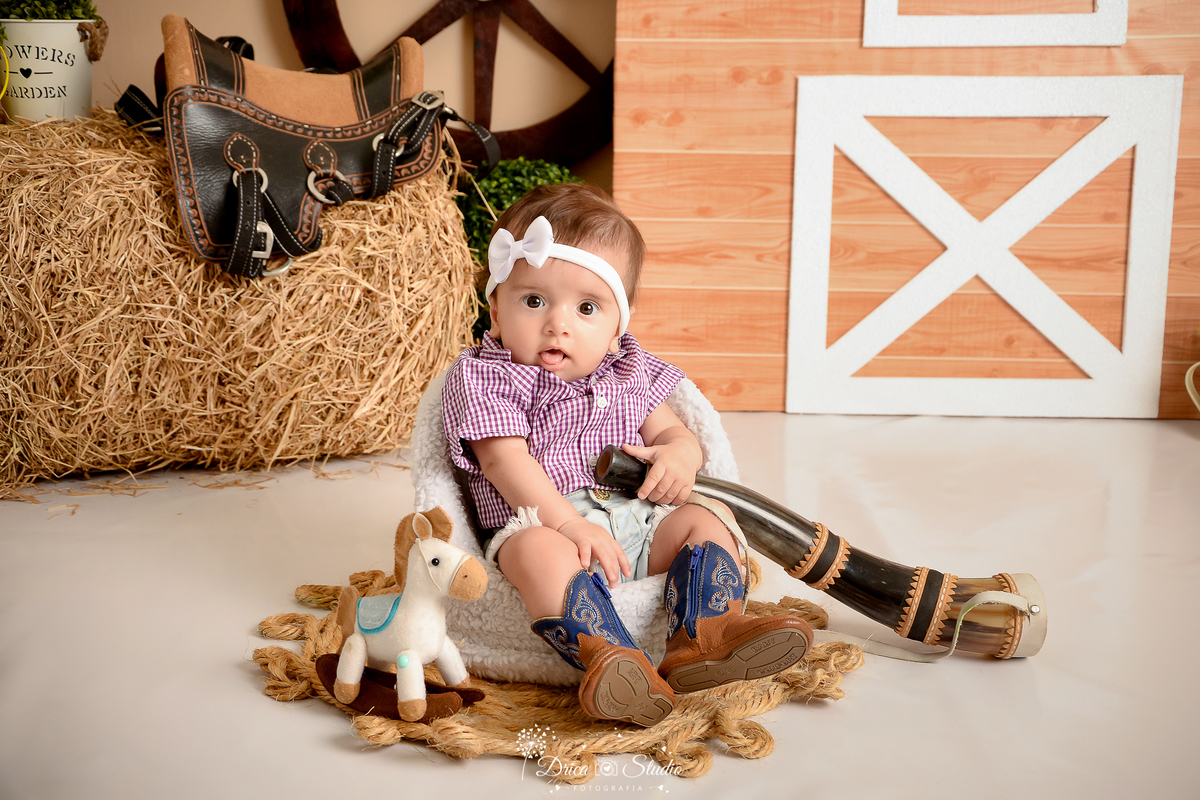 drica studio - ensaio fotografico infantil - fazendinha - xinguara - berrante - feno - sela - seleiro - bebê sentada em poltrona branca, vestindo camisete e short jeans e bota com detalhe azul - cavalinho de brinquedo - tapete rustico de corda