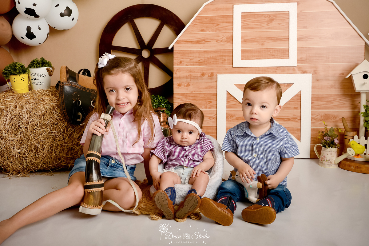 drica studio - ensaio fotografico infantil - fazendinha - xinguara - seleiro - feno - três crianças sentadas no chão, vestindo roupa country - sela- balão branco com mancha preta -rodão de carroça - regador rosa com flores - berrante - fundo bege 