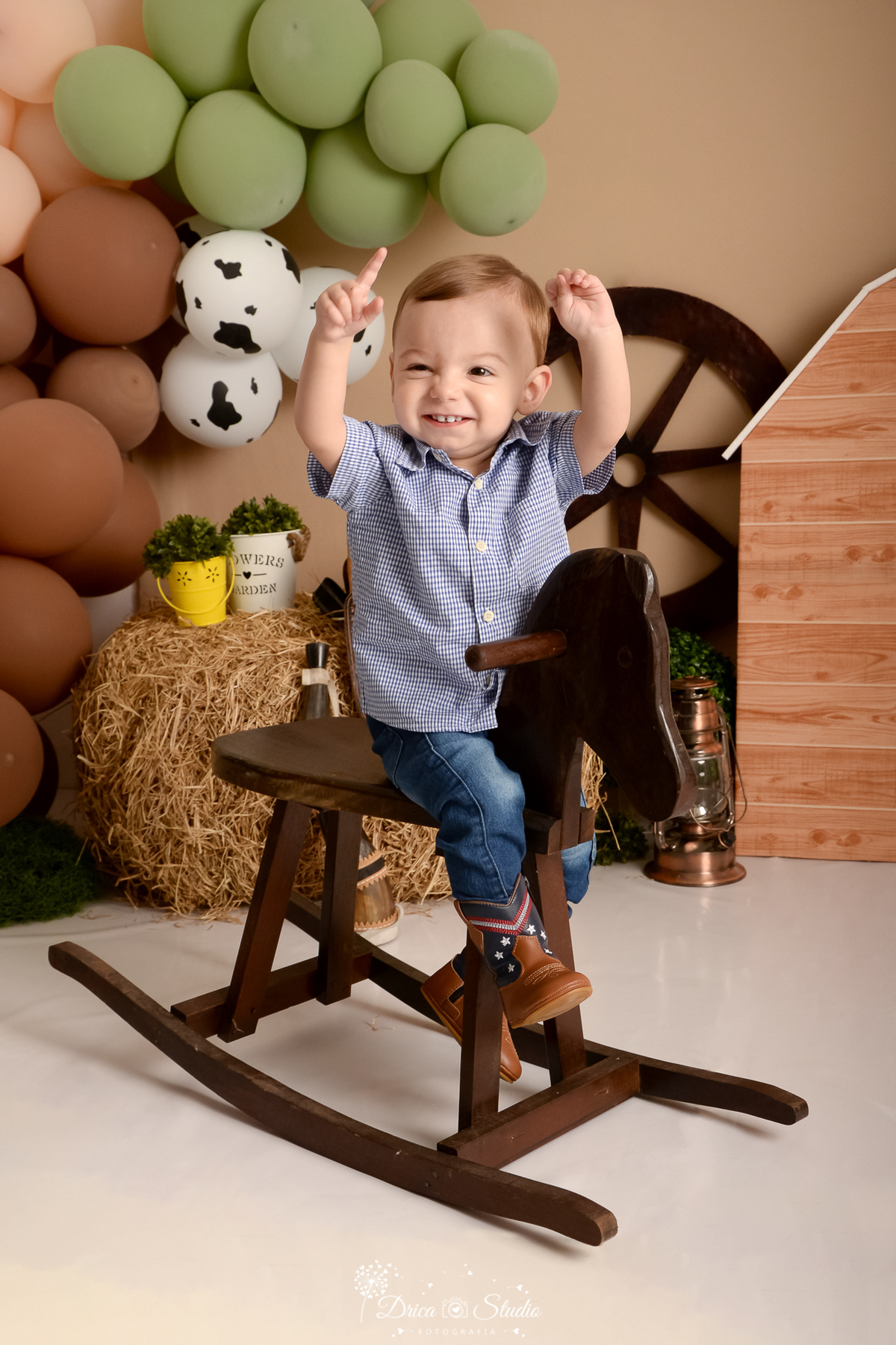drica studio - ensaio fotografico infantil - fazendinha - xinguara - menino sentado em cavalinho de madeira com as mãos erguidas - camisa xadrez e calça jeans - bota marrom com detalhes azul - lampião - feno - balão marrom - baldinho amarelo com arbustos