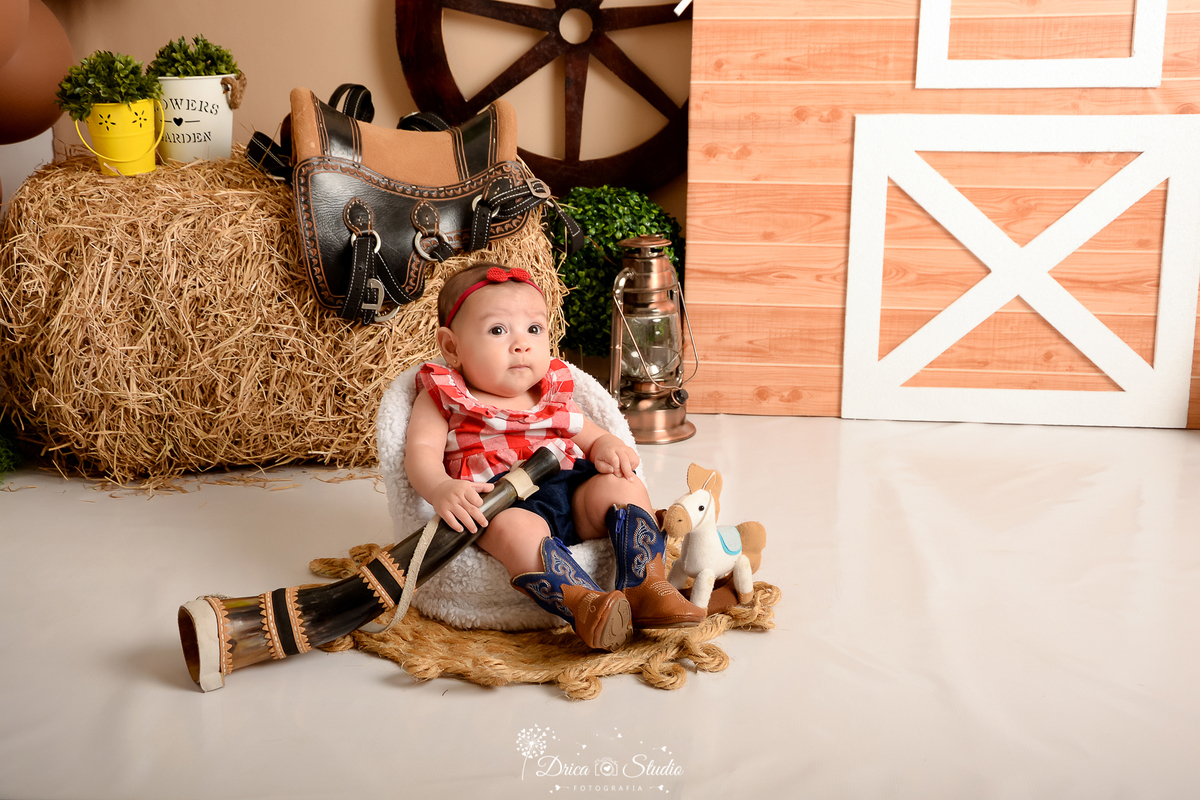 drica studio - ensaio fotografico infantil - fazendinha - xinguara - seleiro - sela - rodão de carroça - bebê sentada em poltrona, vestindo blusa xadrez, short jean escuro e bota marrom com detalhe azul, segurando um berrante - lampiaõ -  tapate rustico