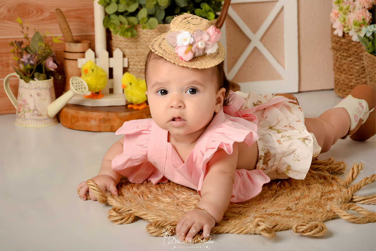 drica studio - ensaio fotografico infantil - fazendinha - xinguara - bebê deitada sobre tapete rustico de corda, vestindo blusa rosa e short creme, chapéu de palha com flores - regador com flores - pintinho - pilão de madeira - cesto de paha com flores 