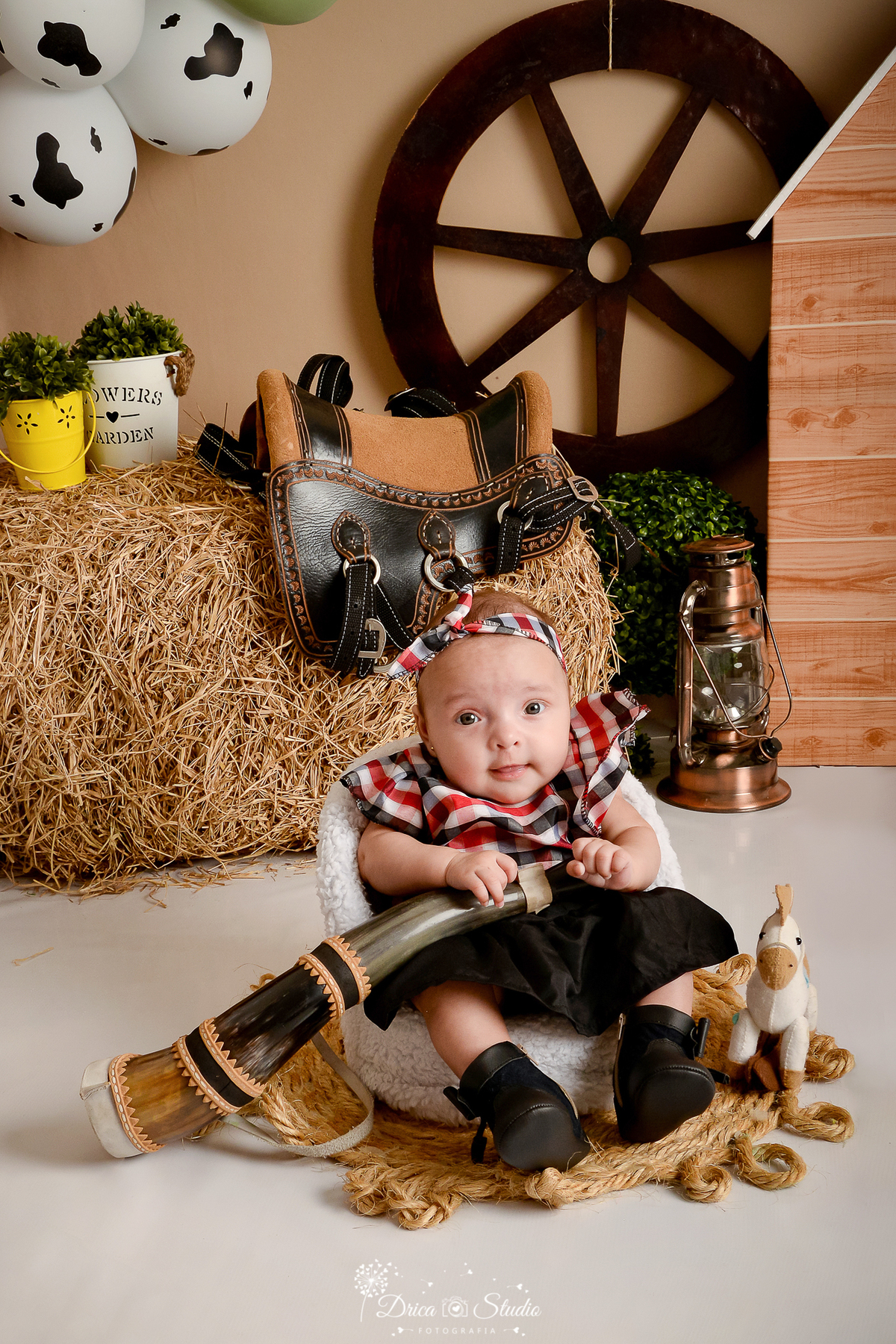 drica studio - ensaio fotografico infantil - fazendinha - xinguara - bebê sentada em poltrona branca, vestindo blusa xadrez e saia preta, segurando berrante - lampião - feno - sela - balde amarelo com arbusto - balde branco com arbusto - rodão de carroça 