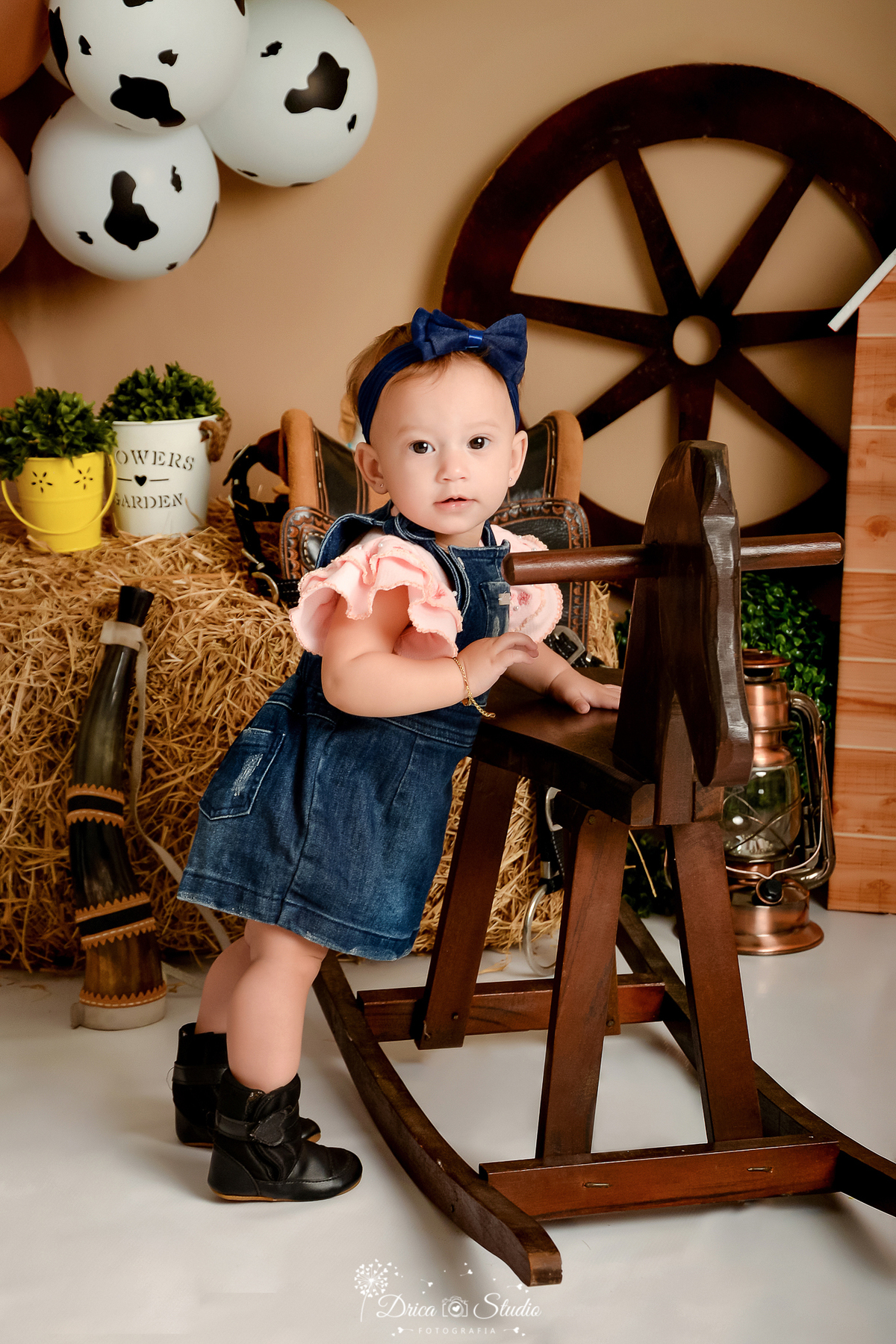 drica studio - ensaio fotografico infantil - fazendinha - xinguara - bebê vestindo jardineira jeans com blusa rosa, em pé ao lado de cavalinho de madeira - lampião - berrante - balde amarelo com arbusto - feno - rodão de carroça - sela - balde branco 