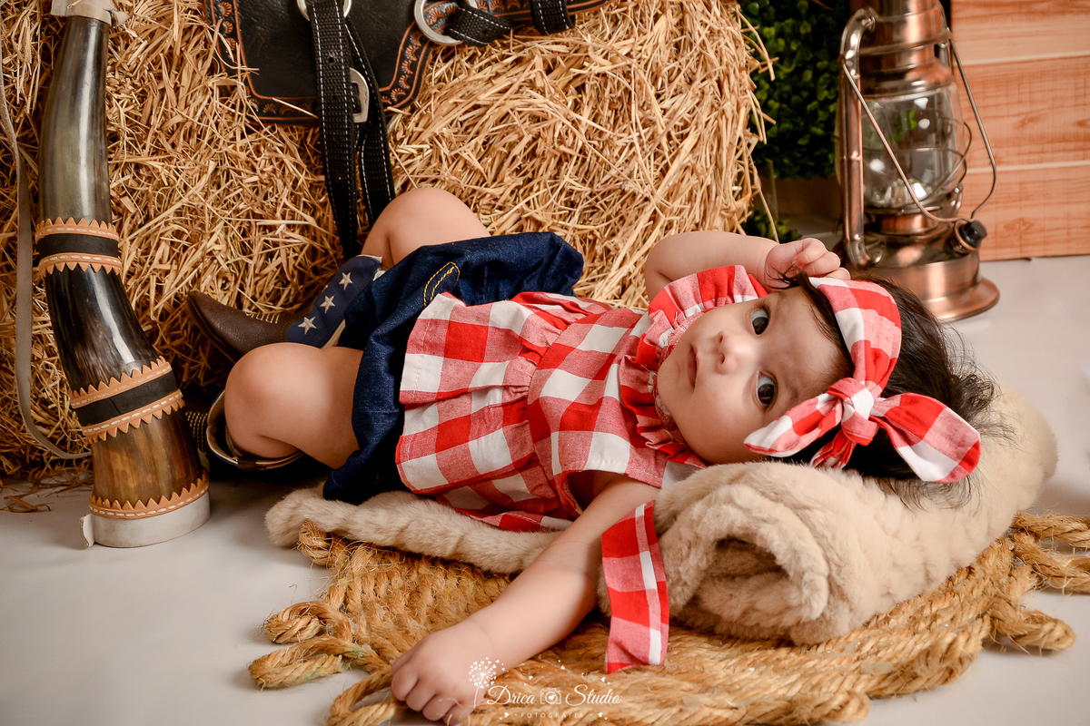 drica studio - ensaio fotografico infantil - fazendinha - xinguara - bebê deitada em manta sobre tapete rustico de corda, vestindo blusa xadrez vermelha com branco - berrante - feno - lampião - sela - seleiro - bota marrom com detalhe azul - short jeans