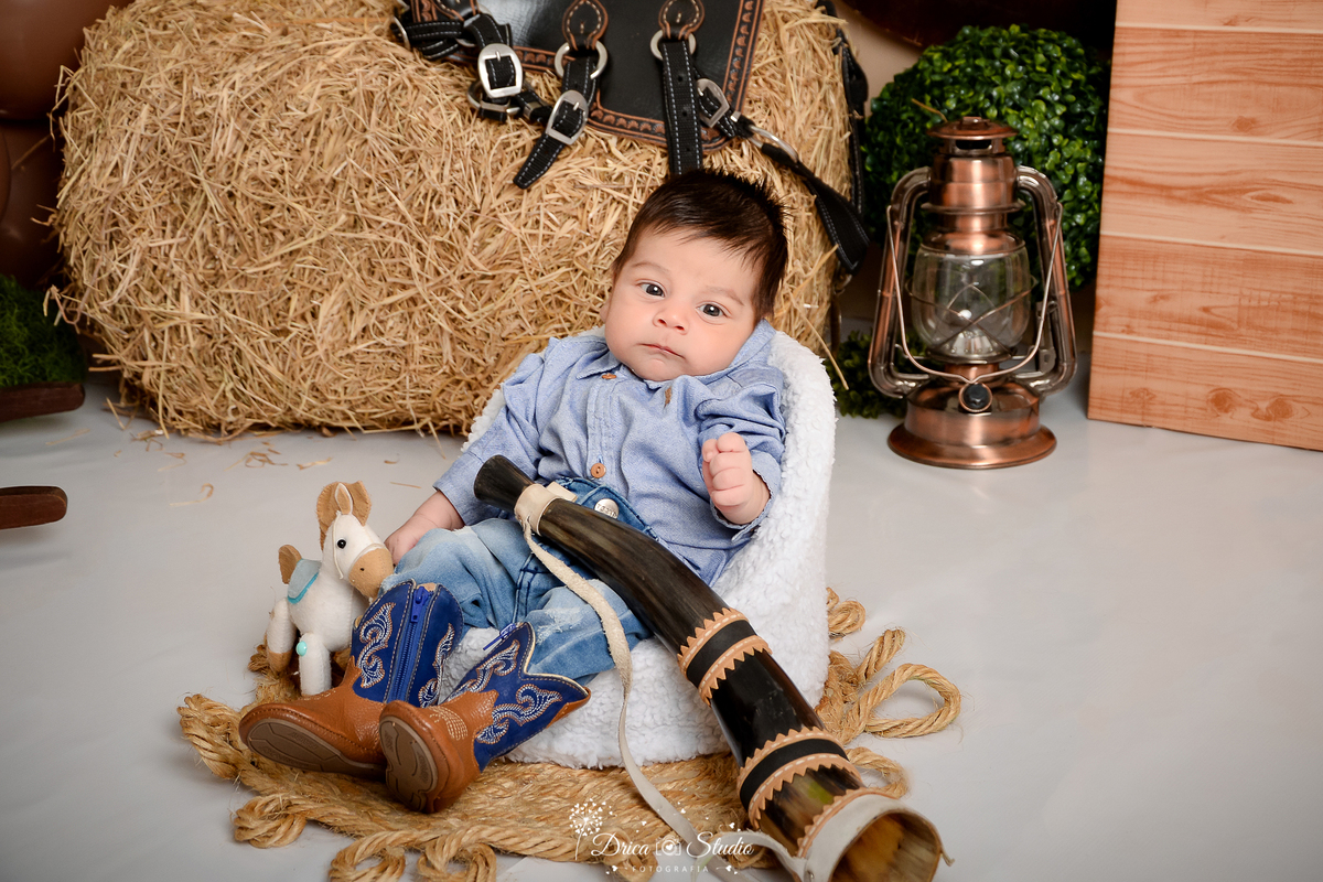 drica studio - ensaio fotografico infantil - fazendinha - xinguara - bebê sentado em poltrona vestindo  calça jeans, camisa jeans azul claro e bota morrom com detalhe azul - lampião - feno - sela - seleiro - arbusto - cavalinho de brinquedo