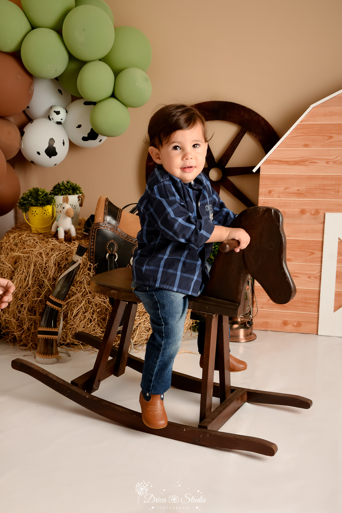 drica studio - ensaio fotografico infantil - fazendinha - xinguara - menino sentado em cavalinho de madeira, vestindo camisa xadrex azul escura com azul claro, calça jeans e bora morrom - berrante - rodão de carroça - sela - balde com arbustos