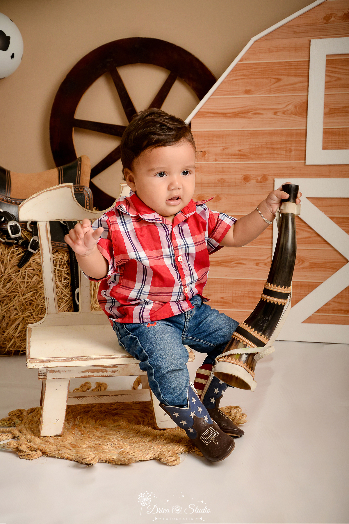 drica studio - ensaio fotografico infantil - fazendinha - xinguara - bebê sentado em cadeira de encosto branca, vestindo camisa vermelha xadrez, calça jeans e bota marrom com detalhe azul, segurando berrante - seleiro - feno - sela - rodão de carroça 