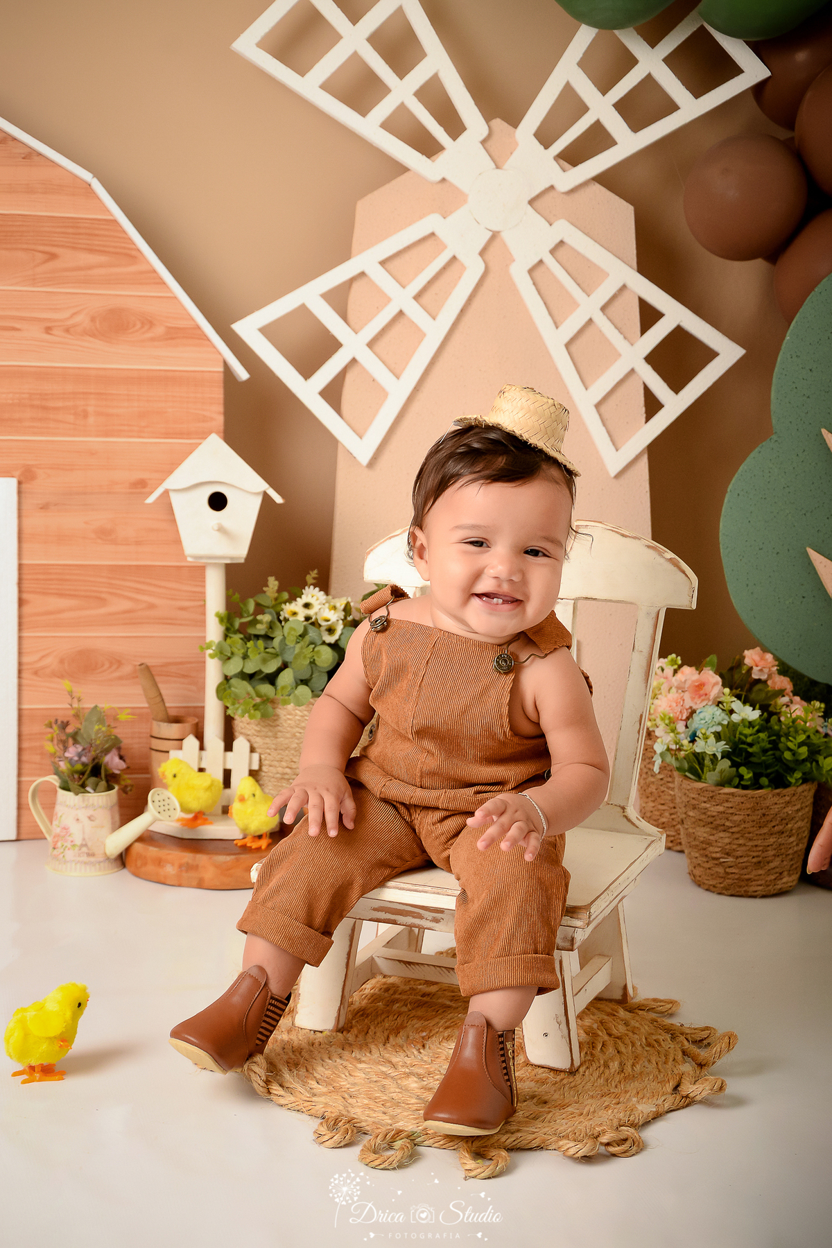 drica studio - ensaio fotografico infantil - fazendinha - xinguara - bebê sorridente vestindo jardineira marrom, bota marrom e chapéu de palha - moinho de vento - cestos com flores - casa de passarinho - regador com flores dentro - pintinho amarelo