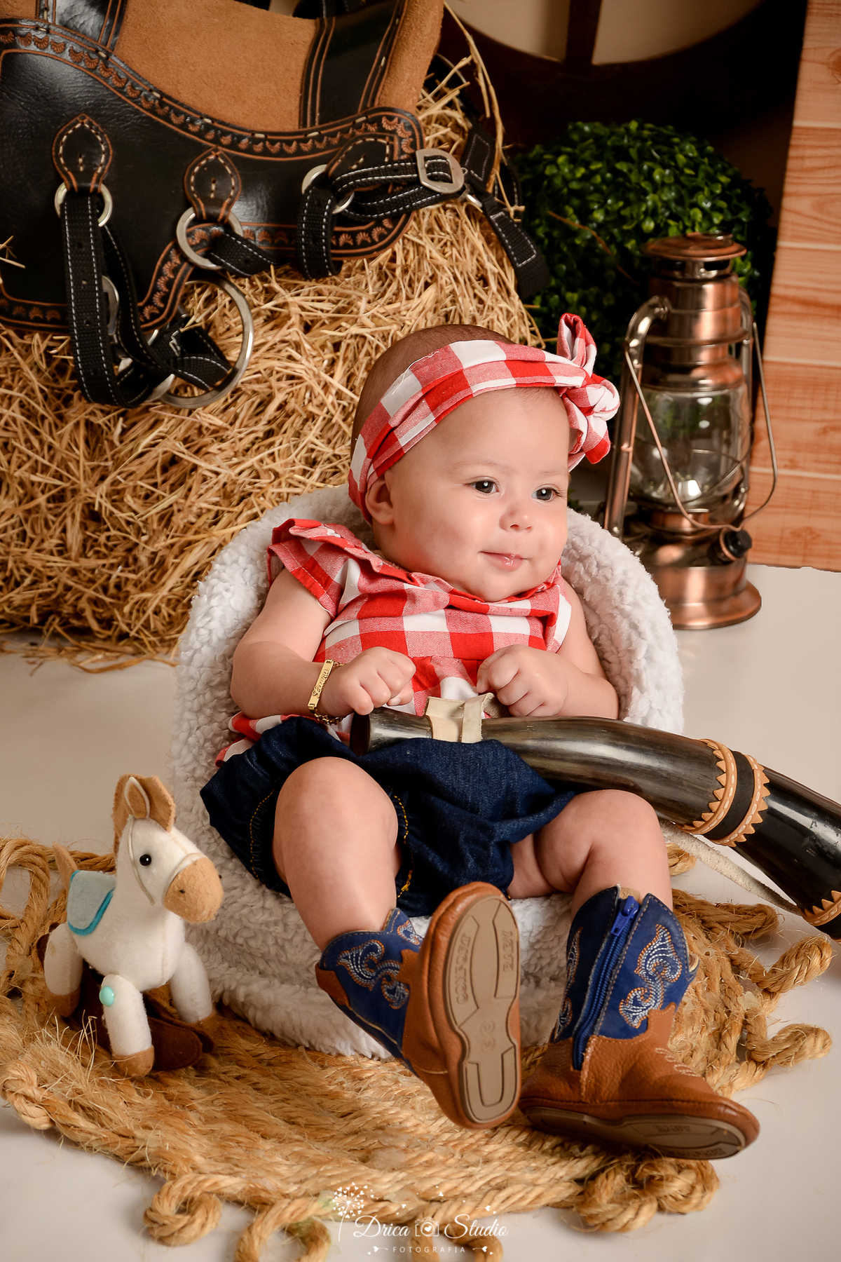 drica studio - ensaio fotografico infantil - fazendinha - xinguara - bebê sentada em poltrona branca, vestindo blusa xadrez vermelha com branco, faixa na cabeça e bota marrom com detalhe azul, segurando berrante - cavalinho de feltro - feno - lampião
