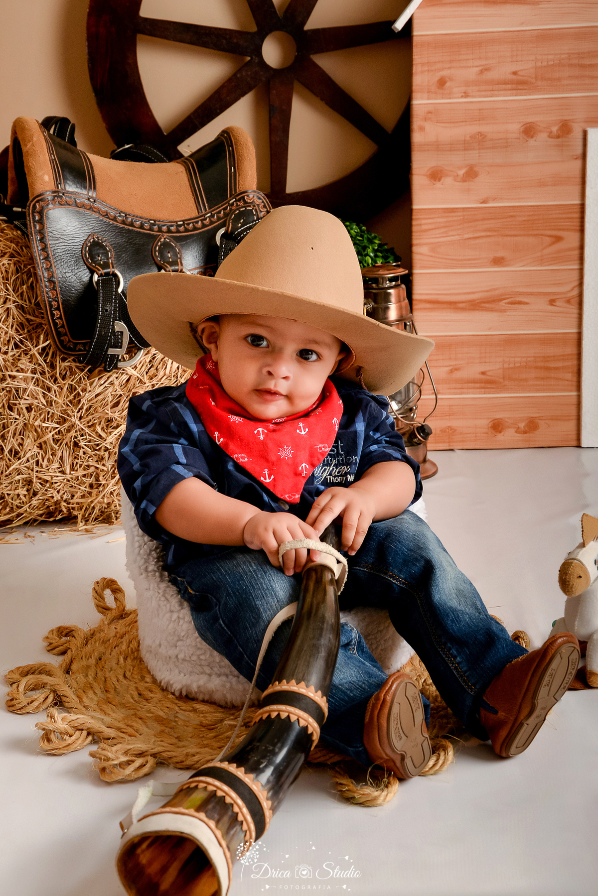 drica studio - ensaio fotografico infantil - fazendinha - xinguara - bebê sentado em poltrona branca, vestindo calça jeans e camisa azul escuro xadrez e lenço vermelho no pescoço, chapéu marrom e bota morrom com detalhe azul - feno - sela - lampião 