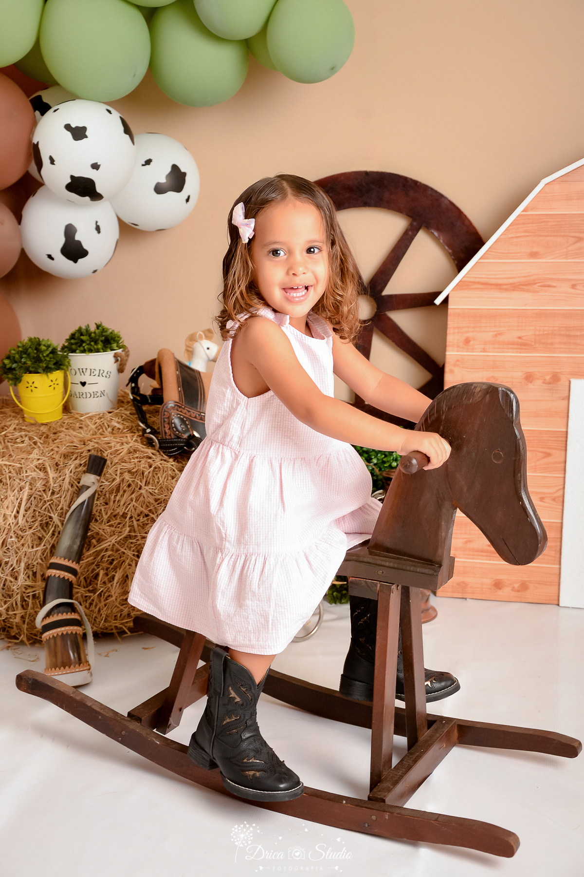 drica studio - ensaio fotografico infantil - fazendinha - xinguara - menina sentada em cavalinho de madeira, usando vestido rosa com bota marrom - berrante - feno - rodão de carroça - seleiro - sela - balde amarelo com arbusto - balão marrom - 