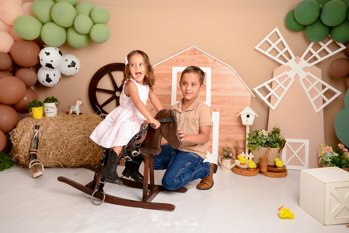 drica studio - ensaio fotografico infantil - fazendinha - xinguara - menina sentada em cavalinho de madeira, usando vestido rosa e bota marrom, menino baixado ao seu lado usando camisa marrom e calça jeans com bota marrom - feno - rodão de carroça - 