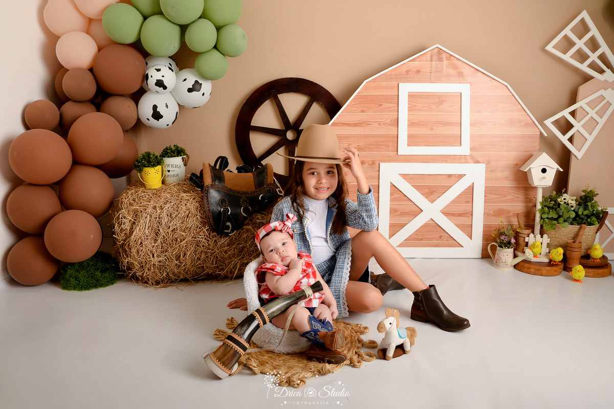 drica studio - ensaio fotografico infantil - fazendinha - xinguara - bebê sentada em poltrona e atras sua irmã sentada no chão- menina usando jaqueta jeans, chapéu e bota marrom - seleiro - feno - balão marrom,creme, verde e branco com manchas preta 