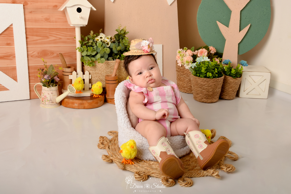 drica studio - ensaio fotografico infantil - fazendinha - xinguara - bebê senta em poltona, vestindo bory xadrez rosa com branco, chapéu de palha com flores e bota morrom com detalhes rosa - cestos de palha com flores - casa de passarinho - regador